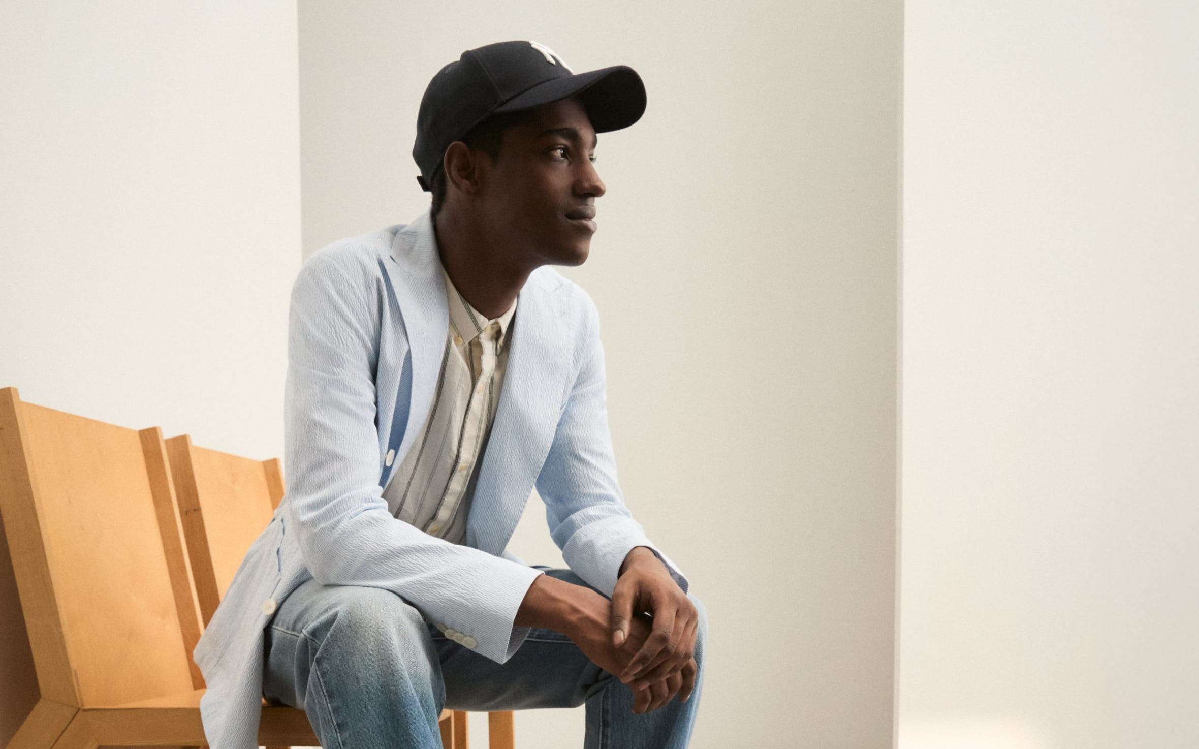 A man wearing a lightweight sport coat, button-down shirt, jeans and ball cap.