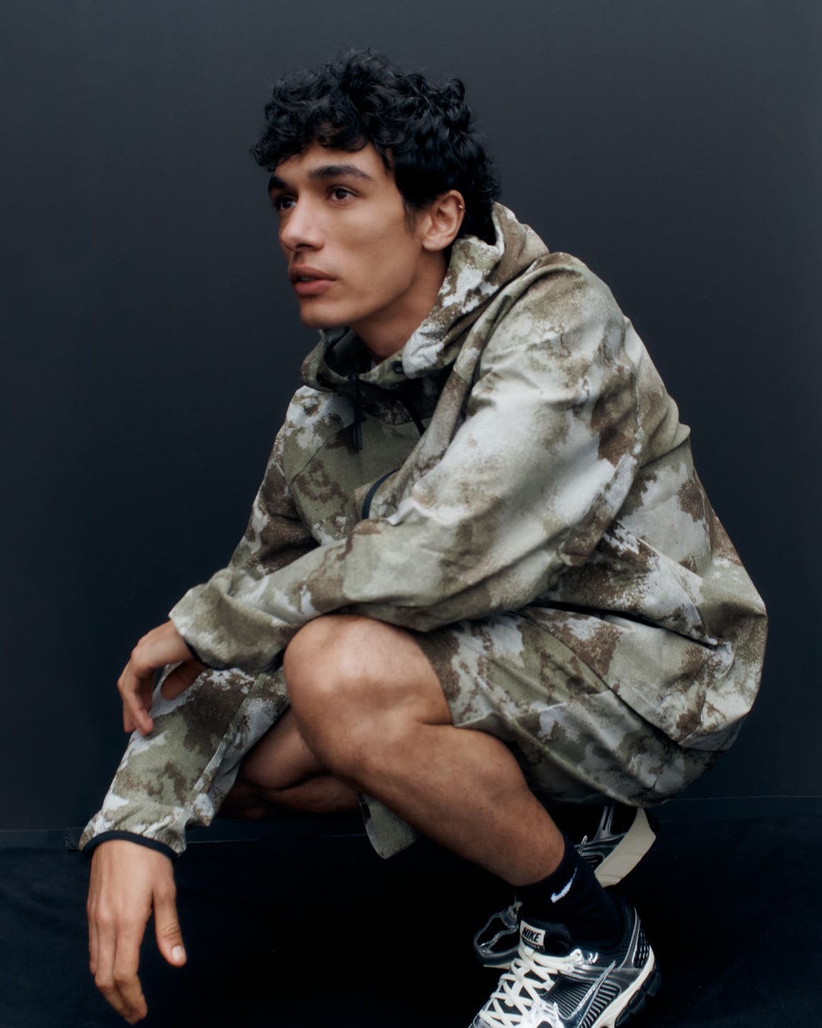 A man wearing a camo-print hoodie and shorts with sneakers.
