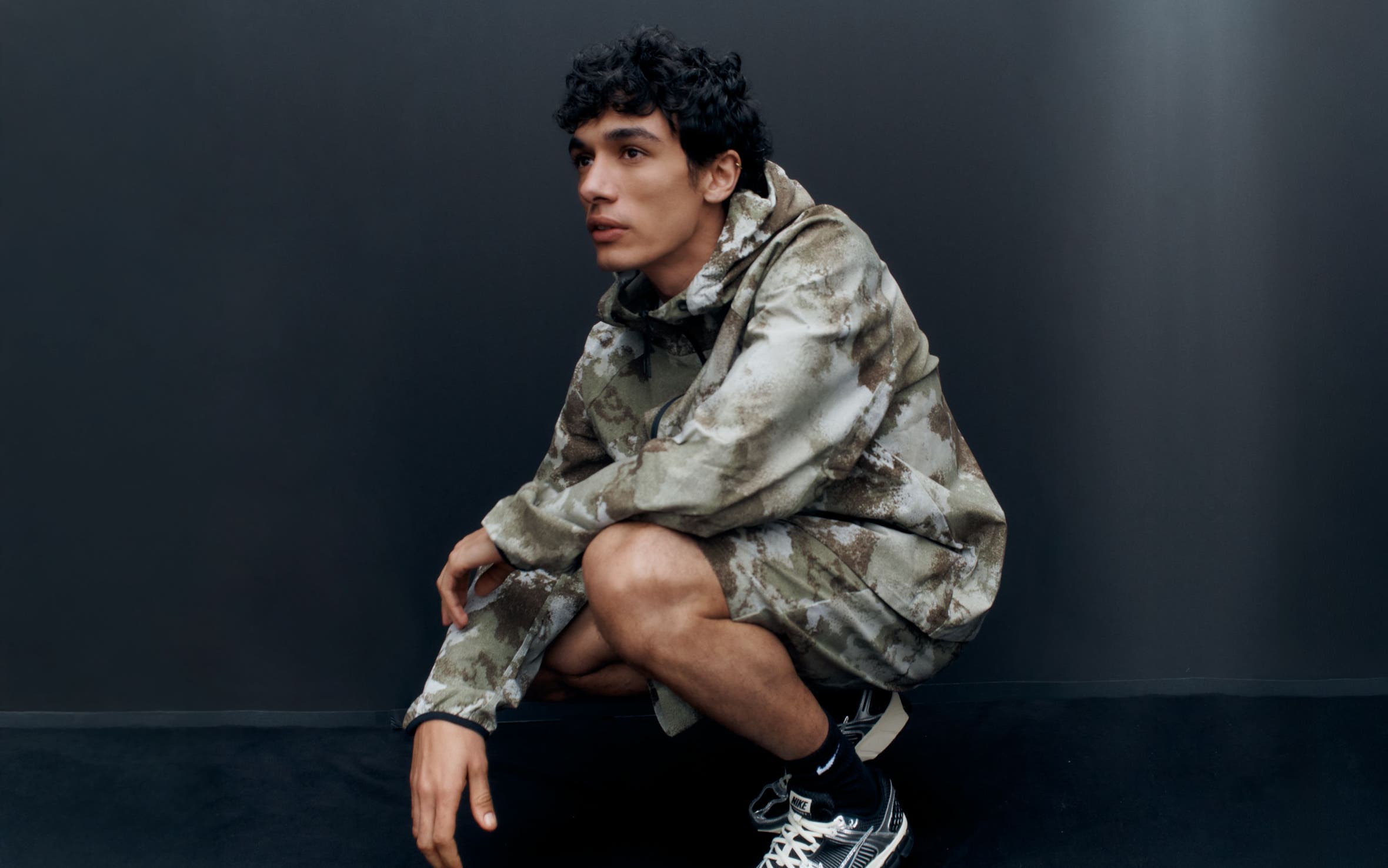 A man wearing a camo-print hoodie and shorts with sneakers.