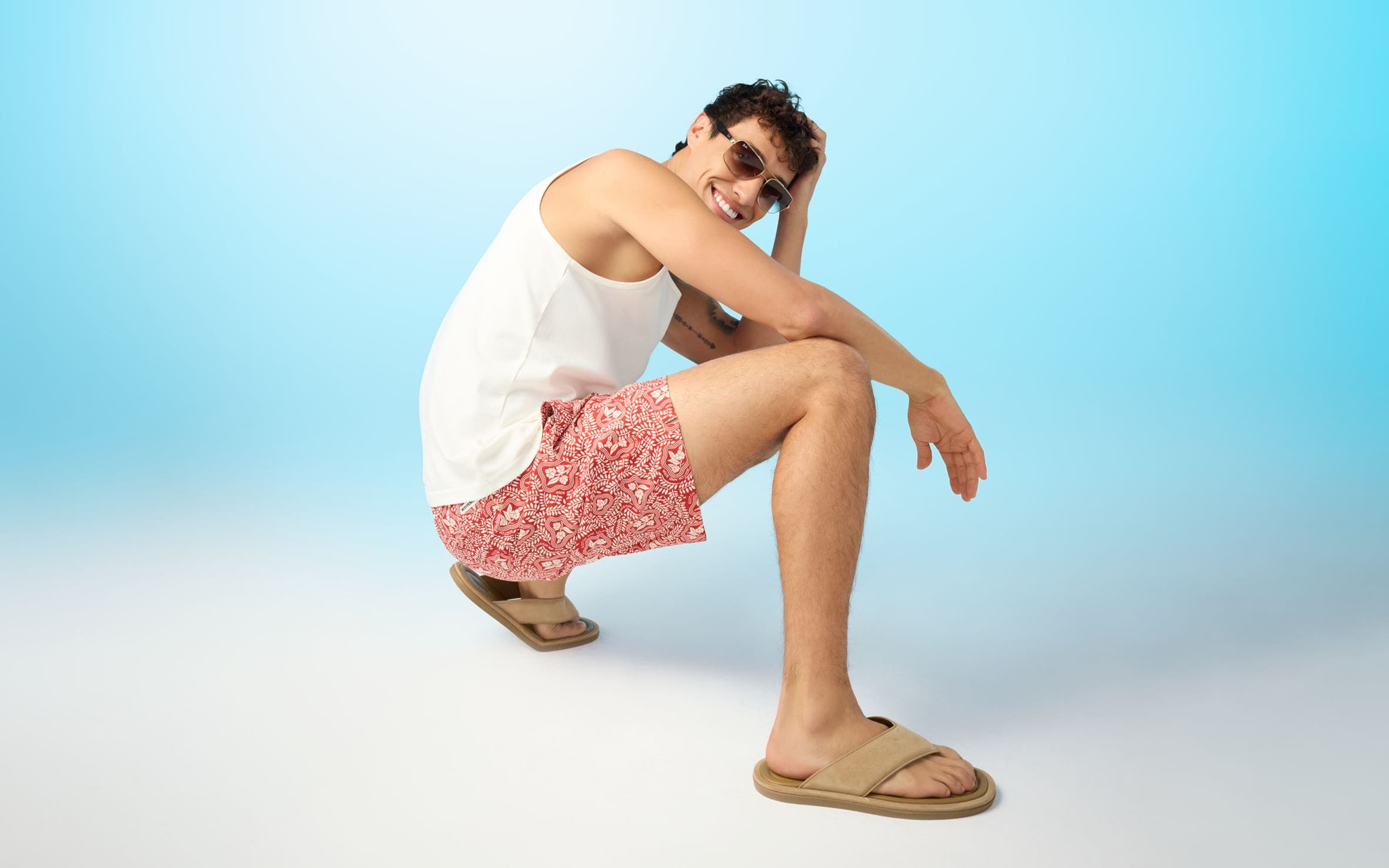 A man wearing a white tank, red floral board shorts, tan sandals and sunglasses.