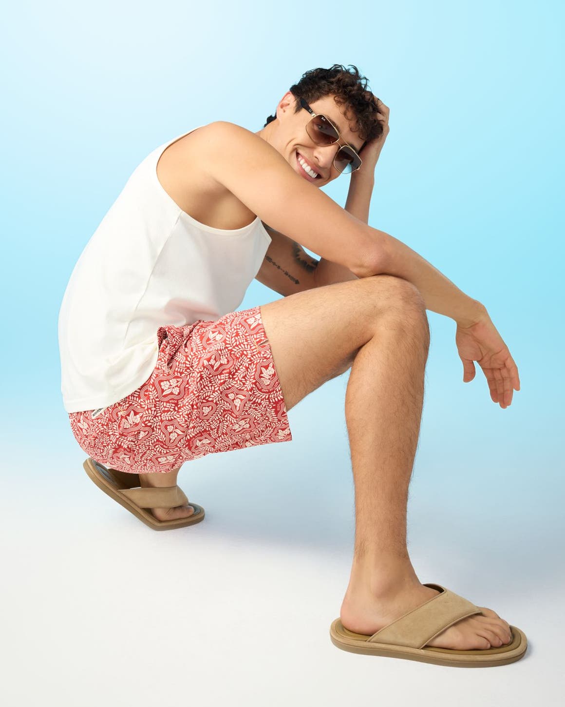 A man wearing a white tank, red floral board shorts, tan sandals and sunglasses.