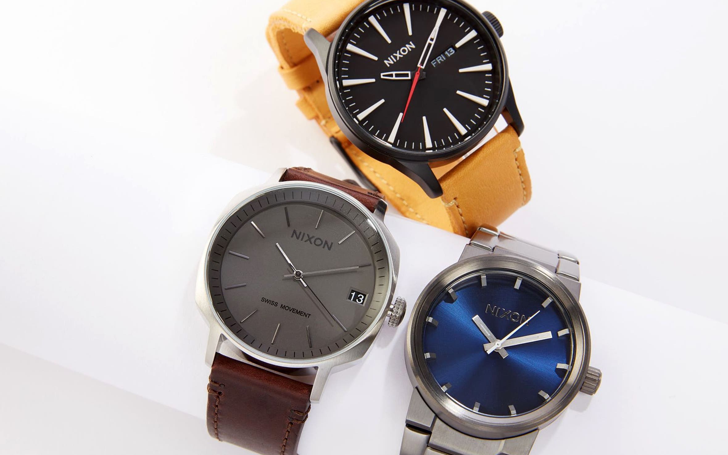 Three Nixon watches: a black dial with tan leather strap, a gray dial with brown leather strap, and a blue dial on a stainless steel bracelet.