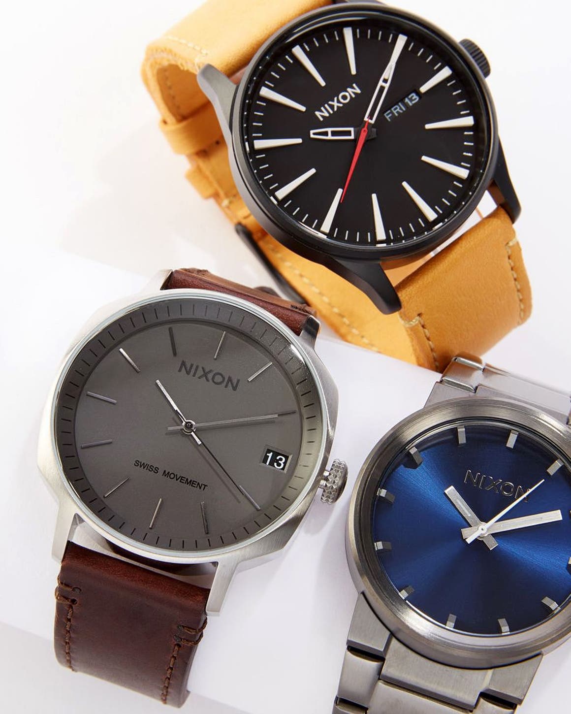 Three Nixon watches: a black dial with tan leather strap, a gray dial with brown leather strap, and a blue dial on a stainless steel bracelet.