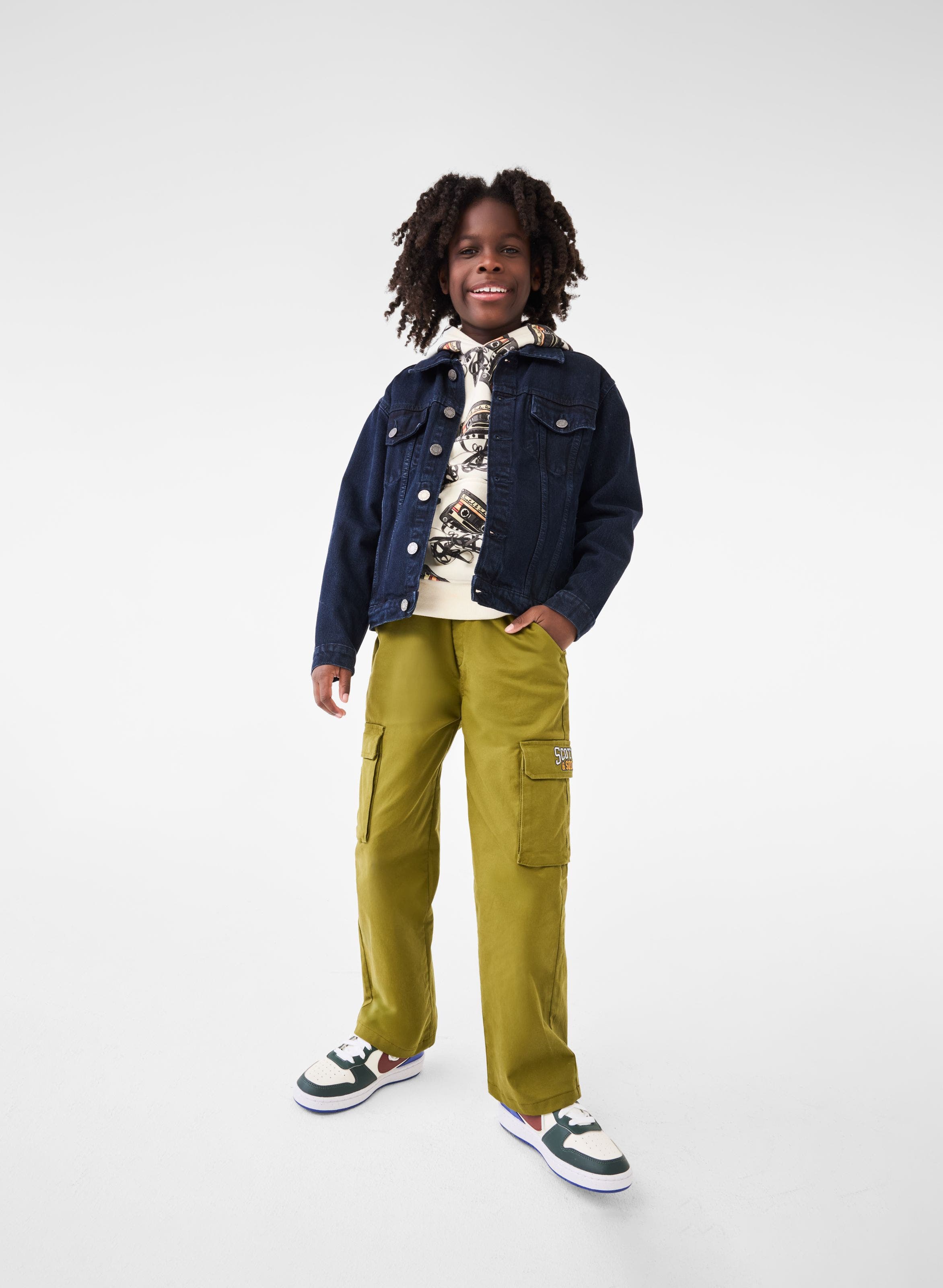 A boy wearing a printed hoodie, a denim jacket, green pants and sneakers.