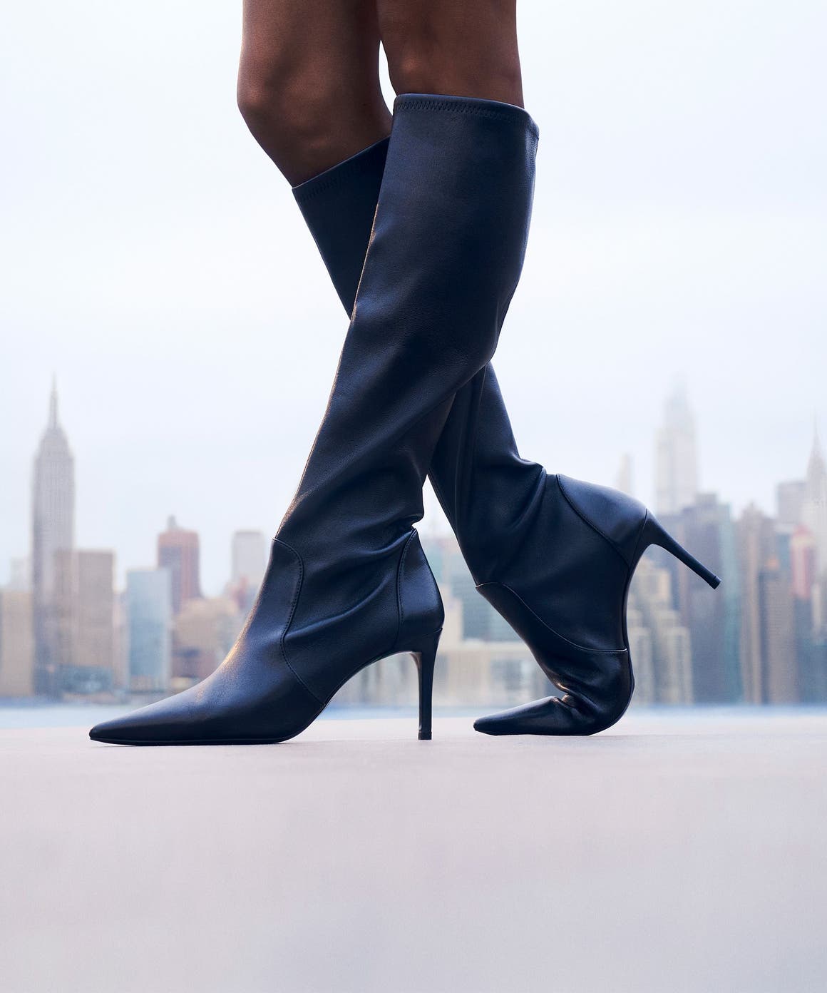 Woman wearing Stuart Weitzman knee-high boots in front of the New York skyline.