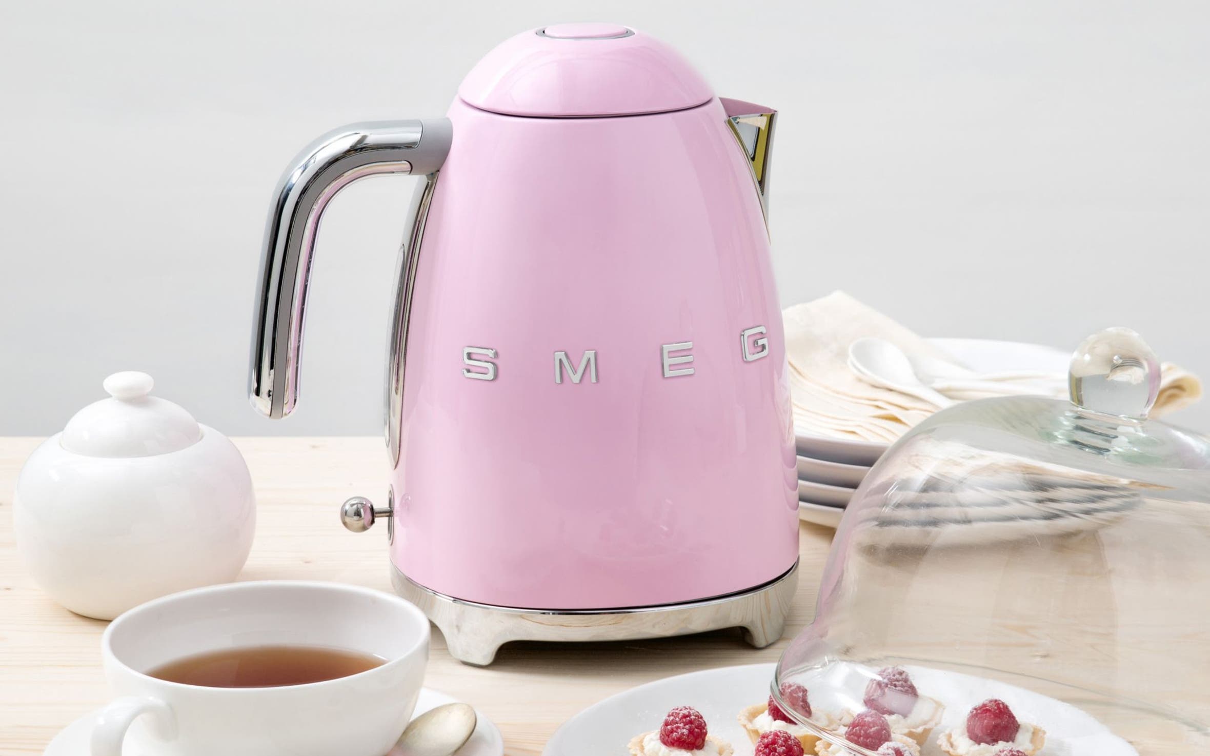 A smeg electric kettle with a tea pot, teacup and plate of pastries.