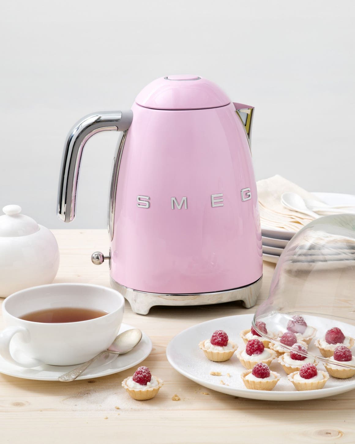A smeg electric kettle with a tea pot, teacup and plate of pastries.