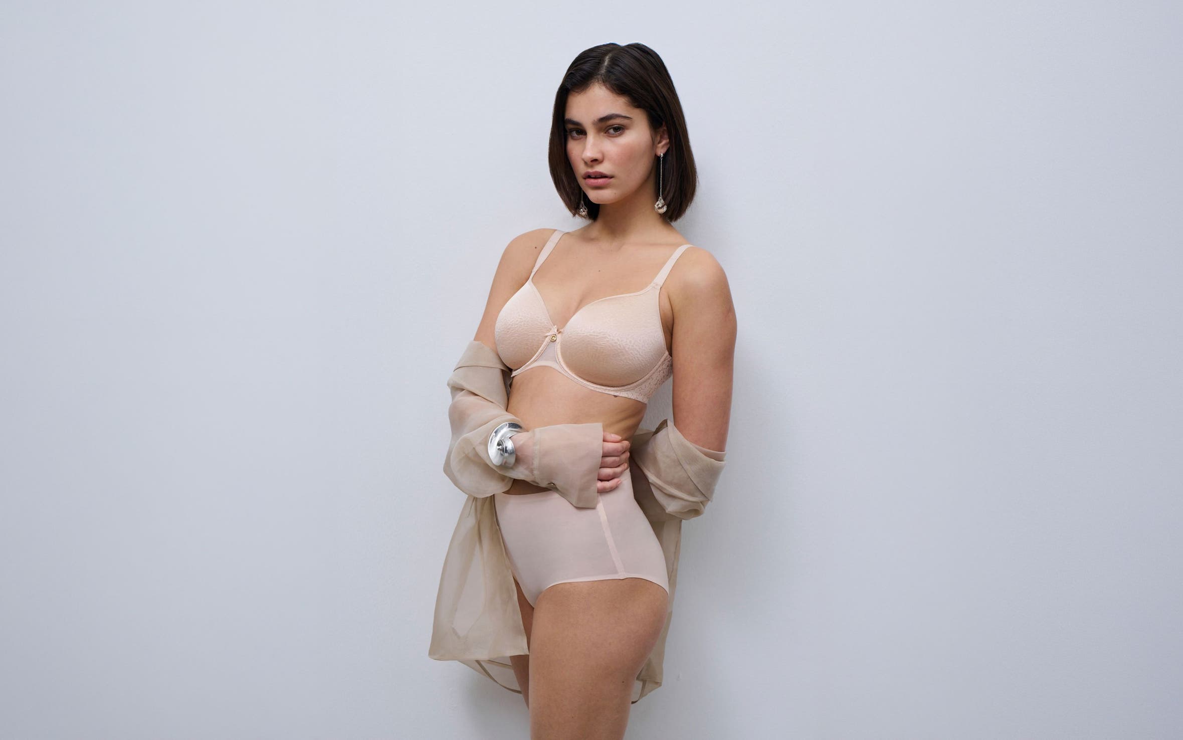 A woman wearing a beige bra and underwear with a sheer shirt.