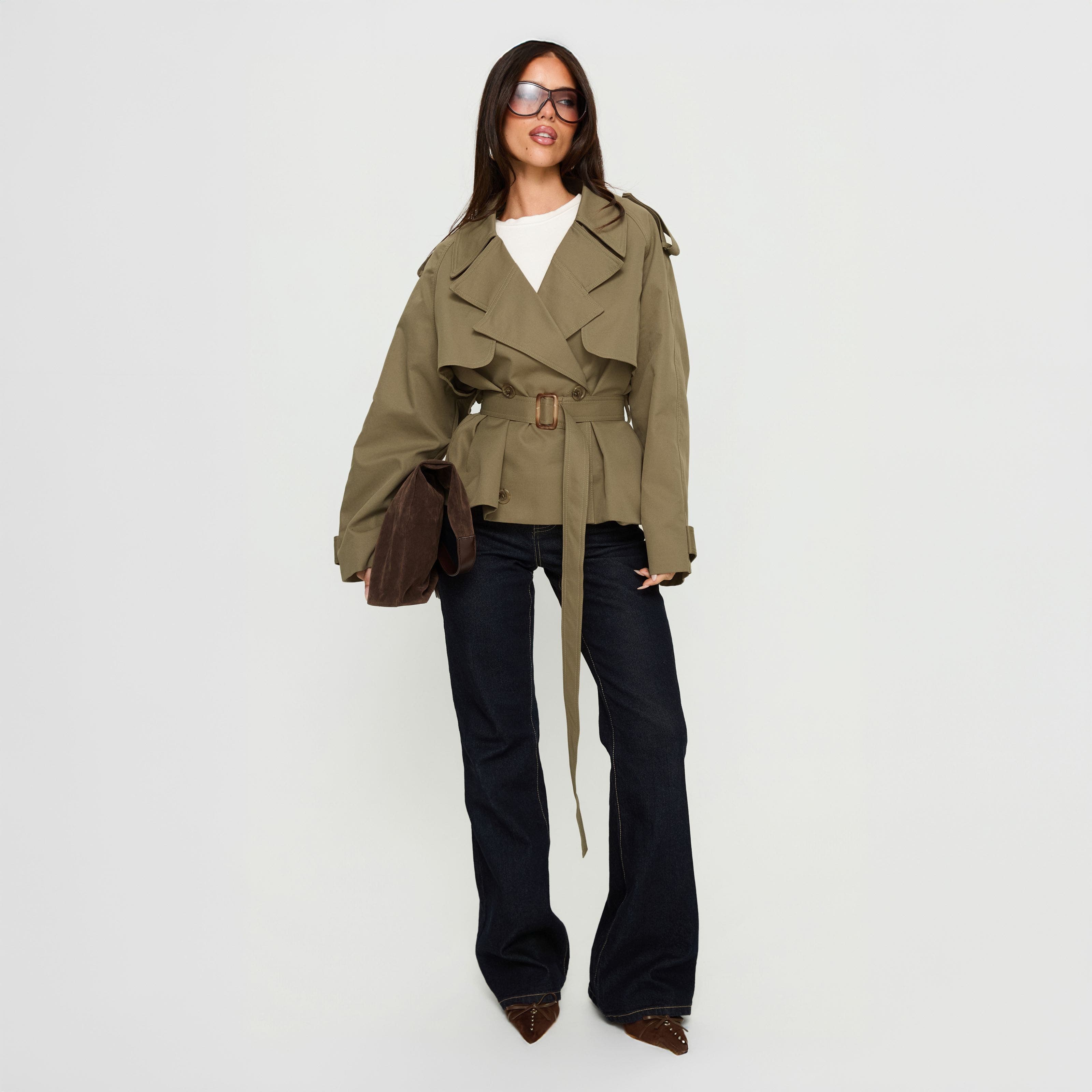 A woman in a cropped army green trench coat over dark-rinse jeans.