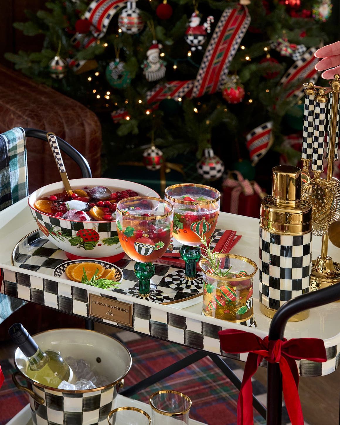 Mackenzie-Childs holiday bar cart styled with a decorative punch bowl, glassware and wine cooler by a Christmas tree.