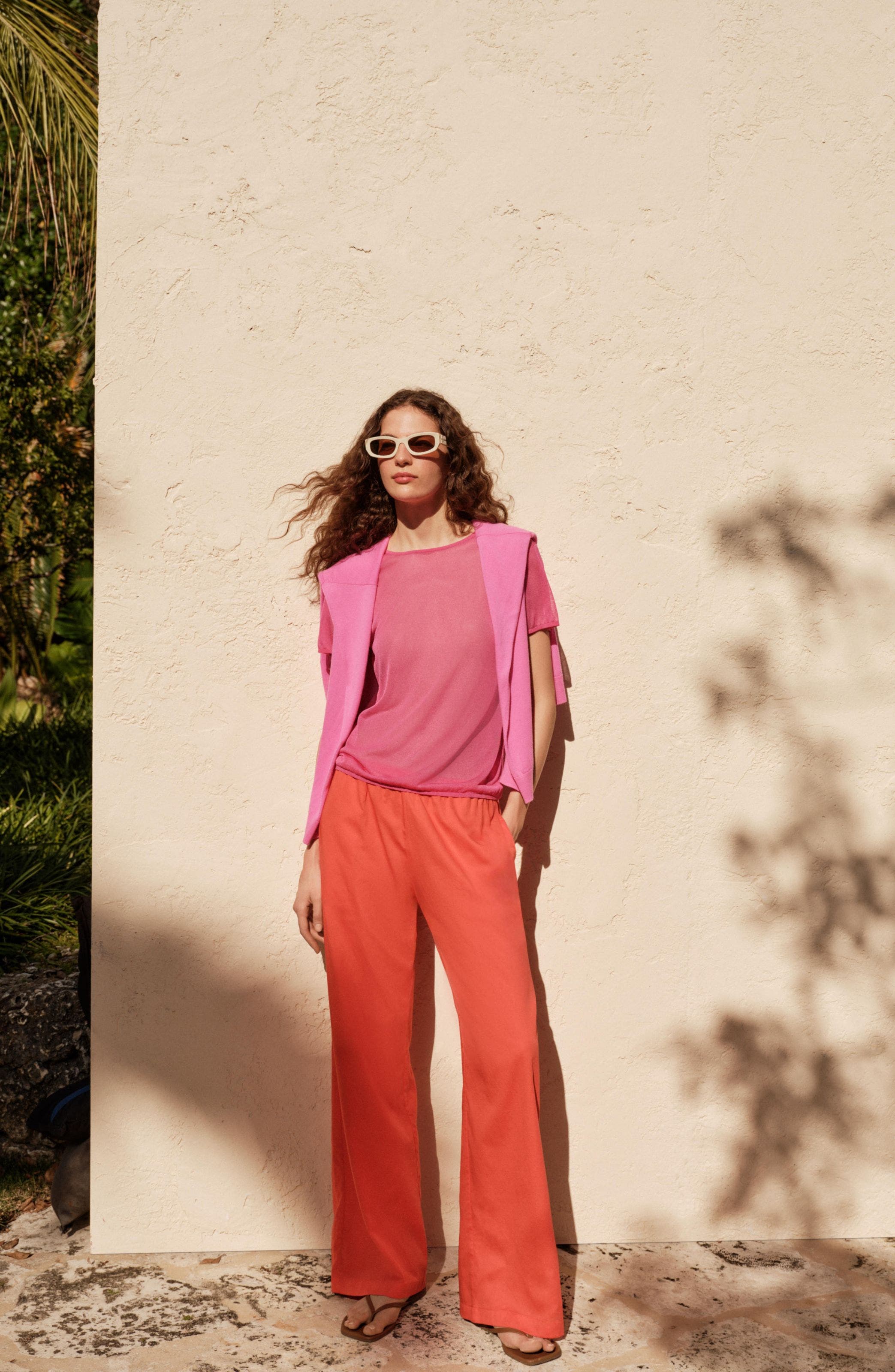 Woman wearing T-shirt, crewneck draped over the shoulders, wide-leg pants, sunglasses and flip-flops.