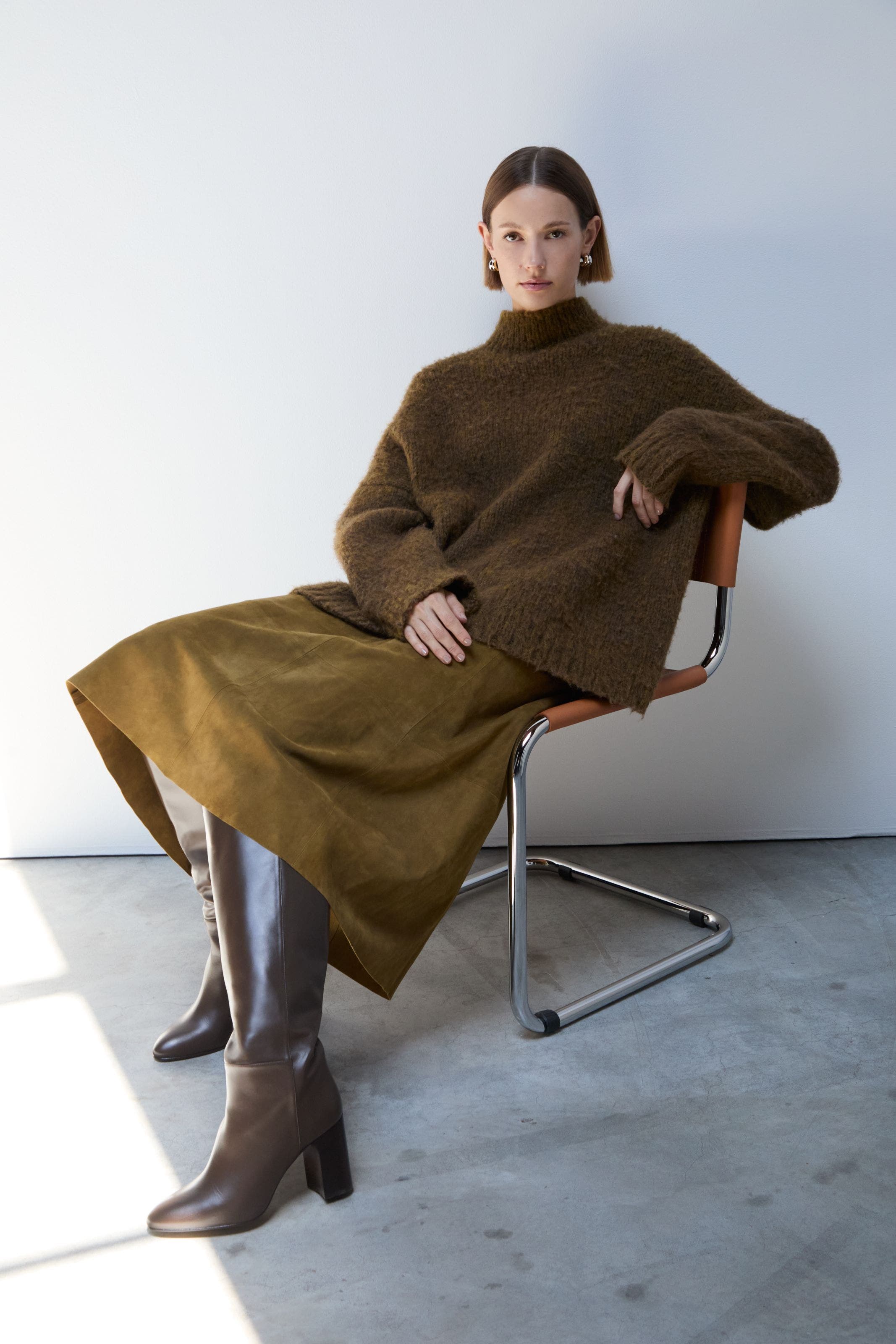 A woman wearing brown leather knee-high boots, an olive green midi skirt and oversized sweater.