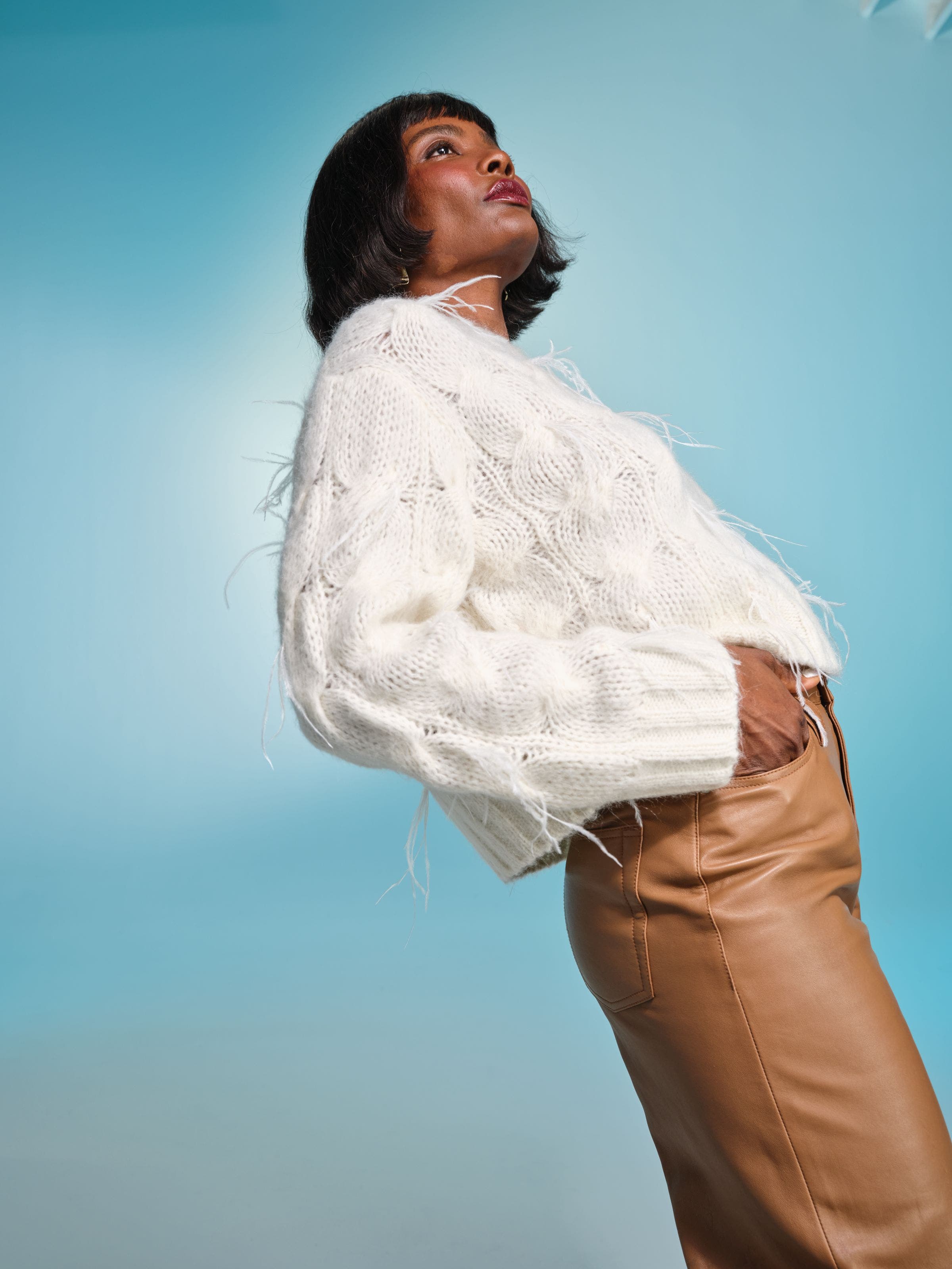 Woman wearing a white fuzzy cable‑knit sweater with subtle fringe details, paired with tan leather pants