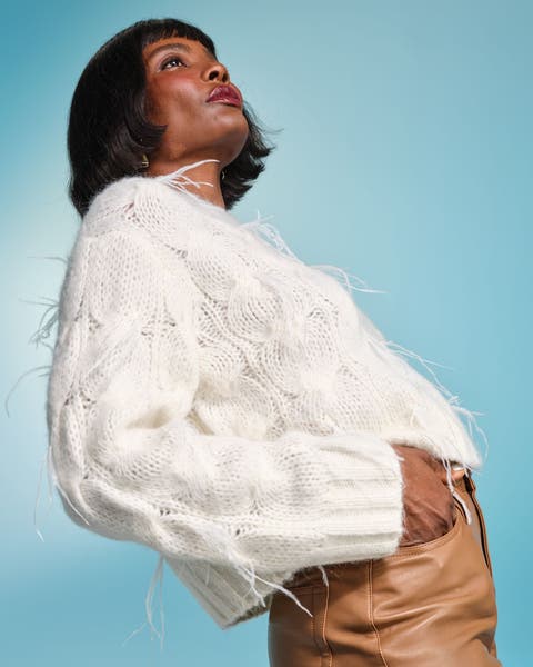 Woman wearing a white fuzzy cable‑knit sweater with subtle fringe details, paired with tan leather pants