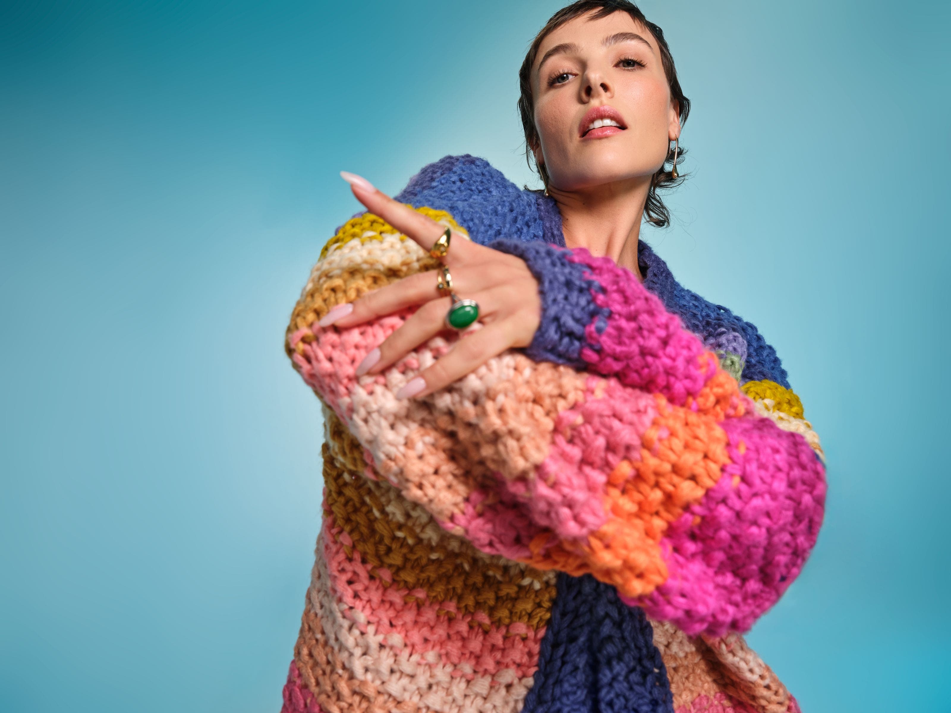 Model wearing an oversized multicolor chunky knit sweater with gold and green statement rings.