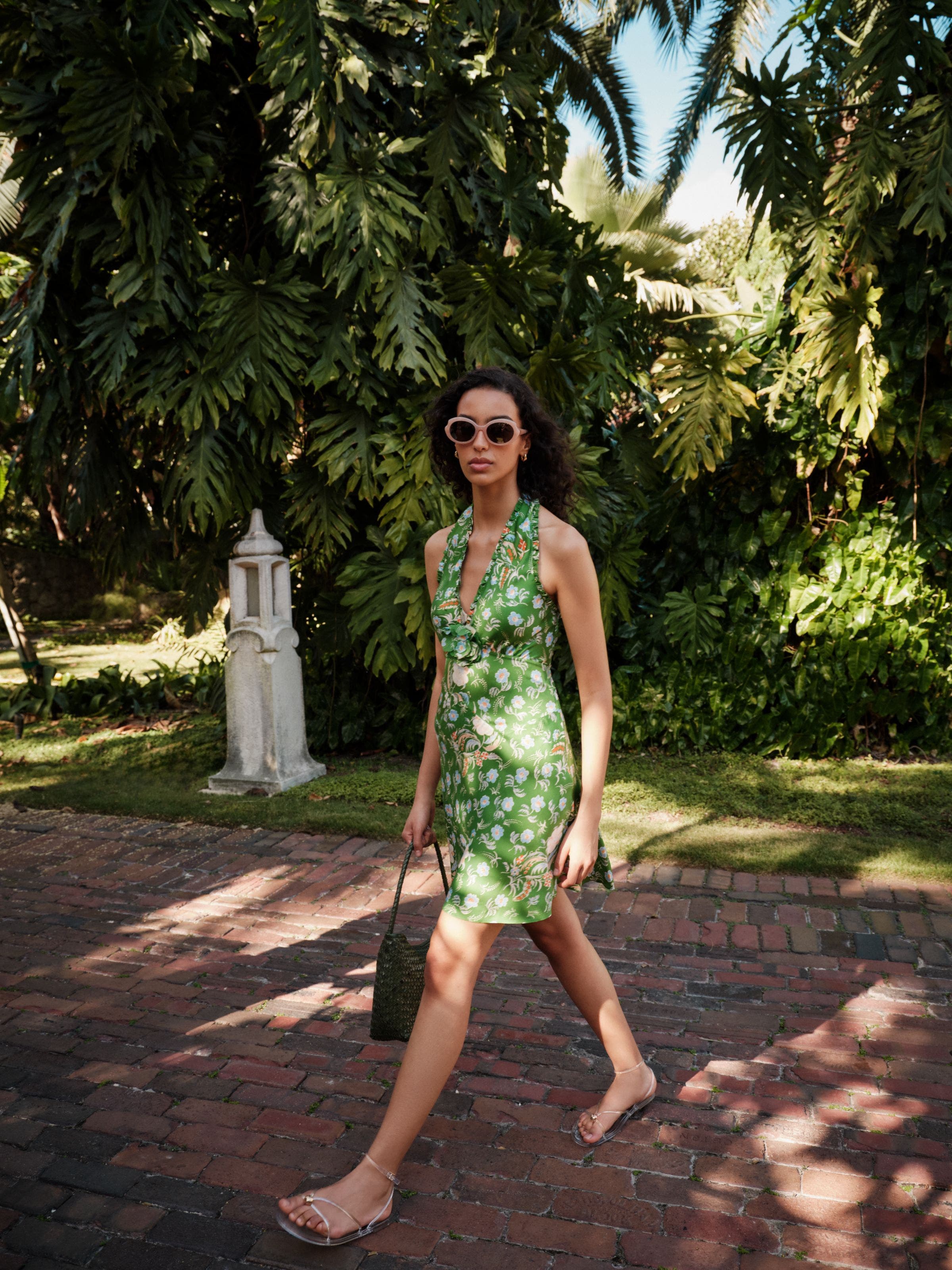 Woman wearing green printed summer dress, sunglasses, sandals and a woven handbag.