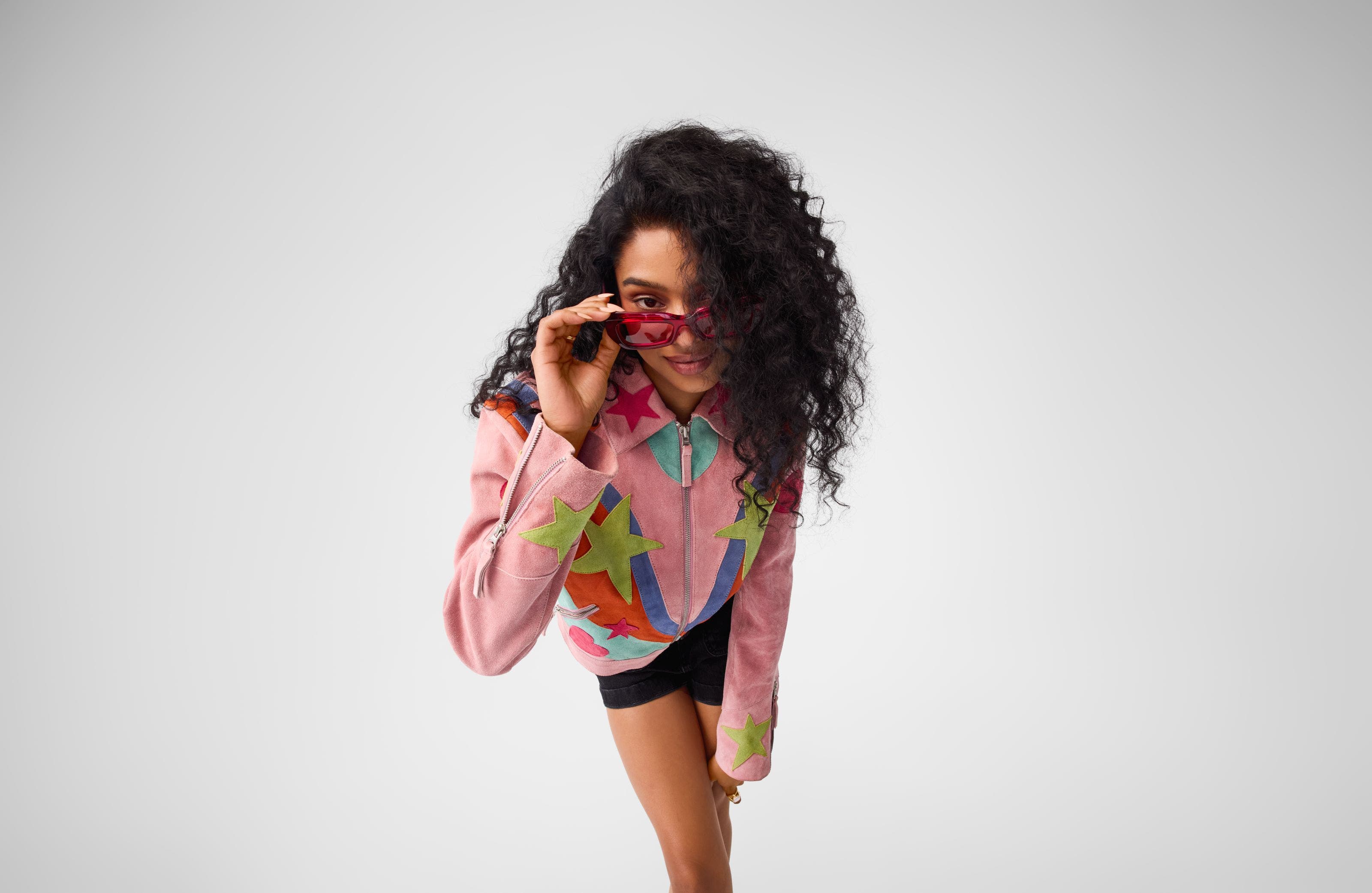 A woman wearing red sunglasses, black shorts and a colorful jacket with star appliques.