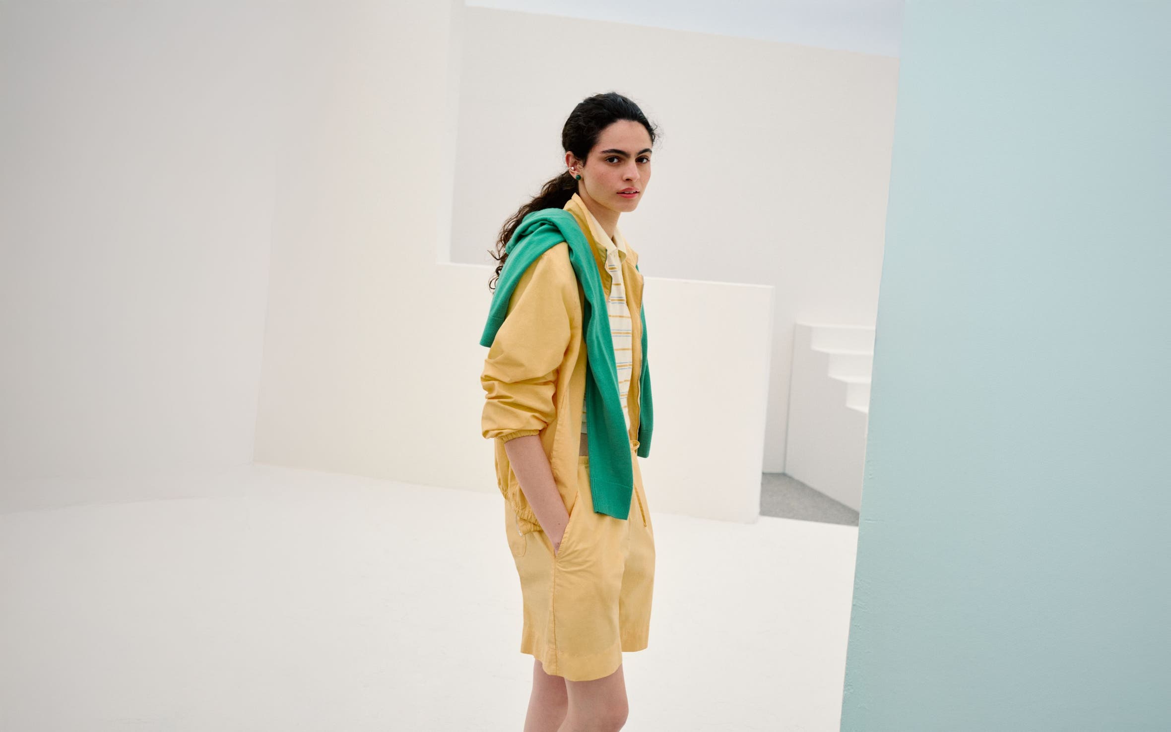 A woman wearing a yellow shirt and shorts over a striped polo top with a green sweater on her shoulders.