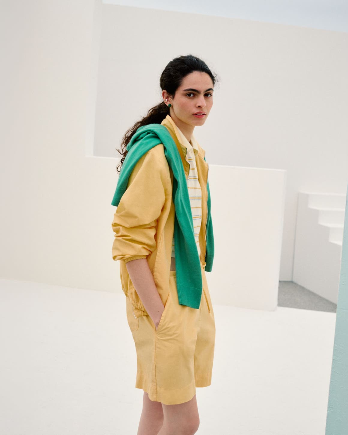 A woman wearing a yellow shirt and shorts over a striped polo top with a green sweater on her shoulders.