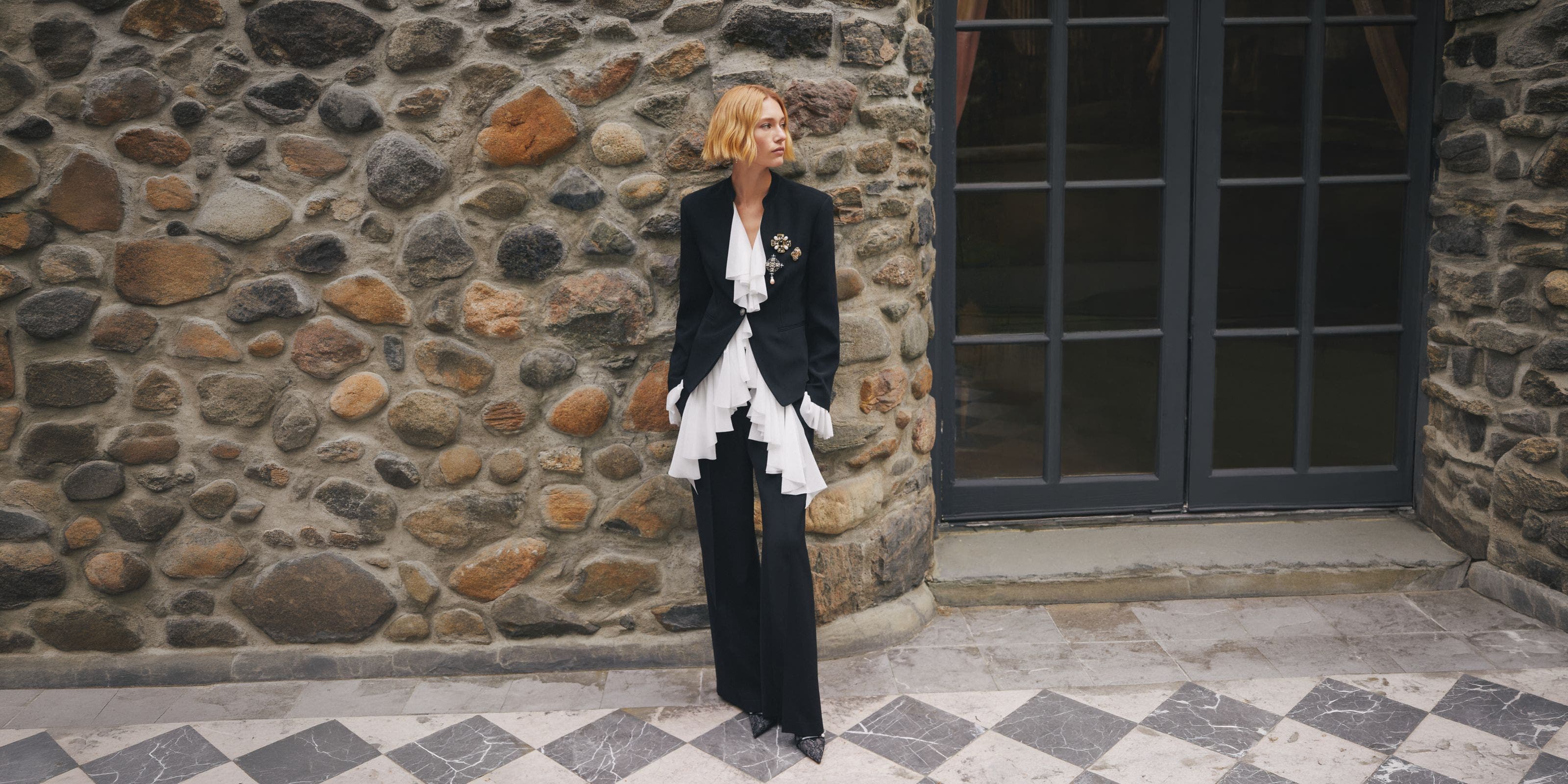 A woman wearing a tailored black suit with layered white ruffles stands against a rustic stone wall. 