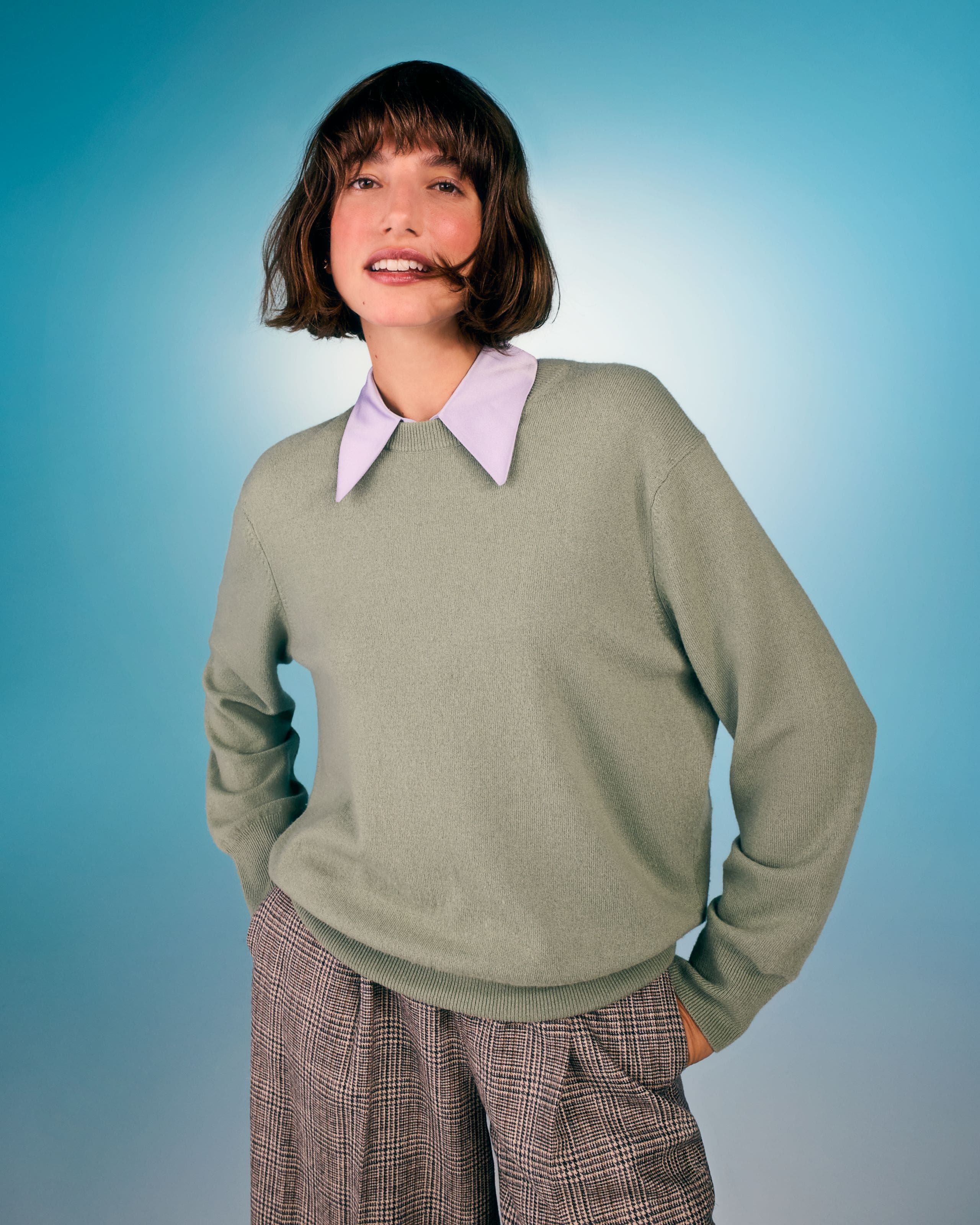 Woman wearing a light sage green sweater over a lavender pointed-collar shirt, paired with plaid trousers.