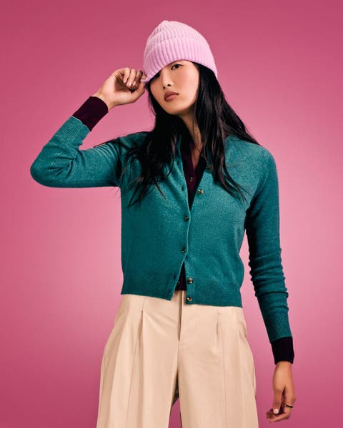 Woman in a teal cardigan, pink beanie, and cream wide-leg trousers.