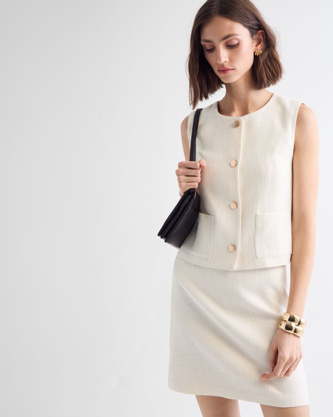 Woman wearing a white vest and skirt with a handbag and jewelry.