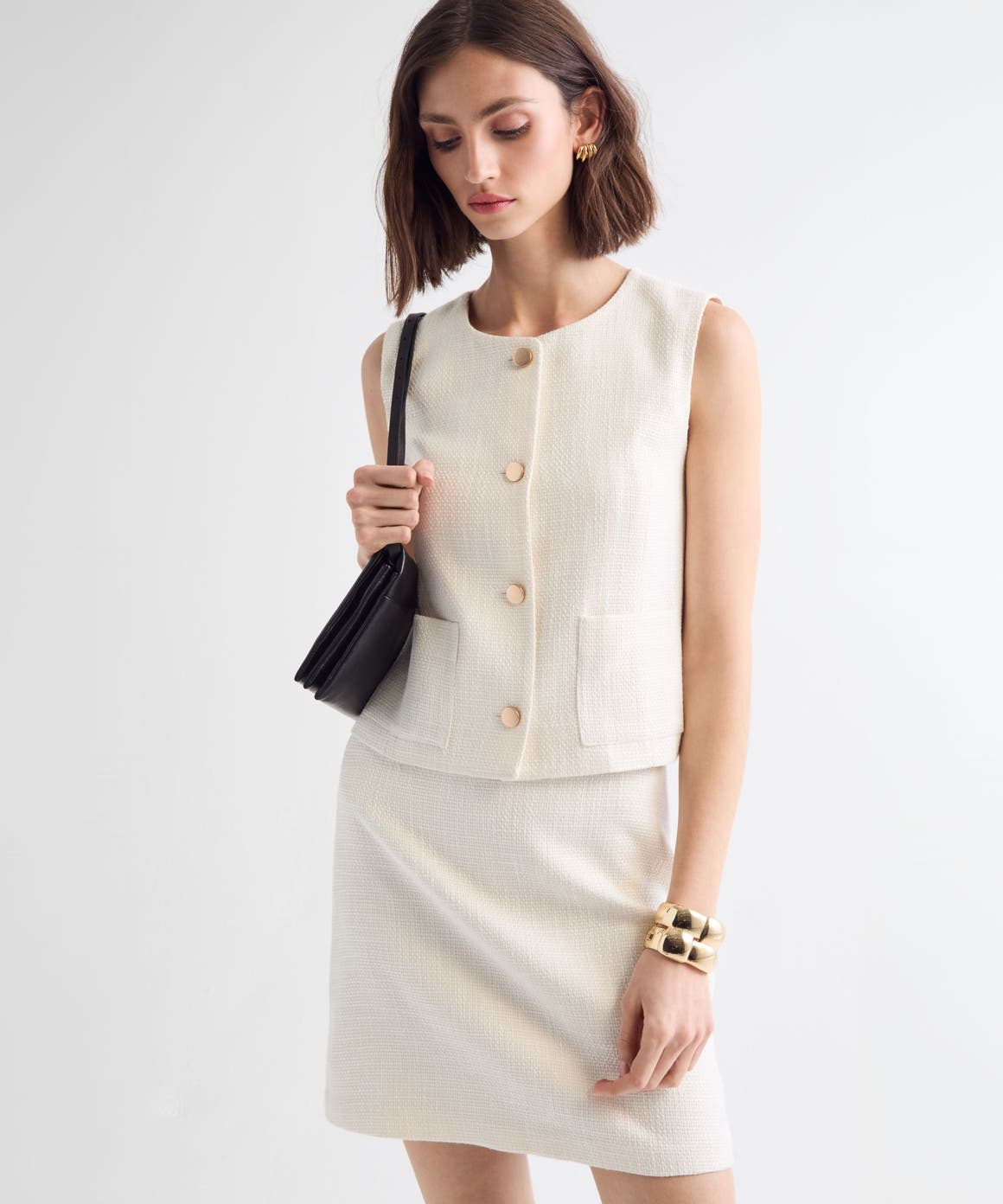 Woman wearing a white vest and skirt with a handbag and jewelry.