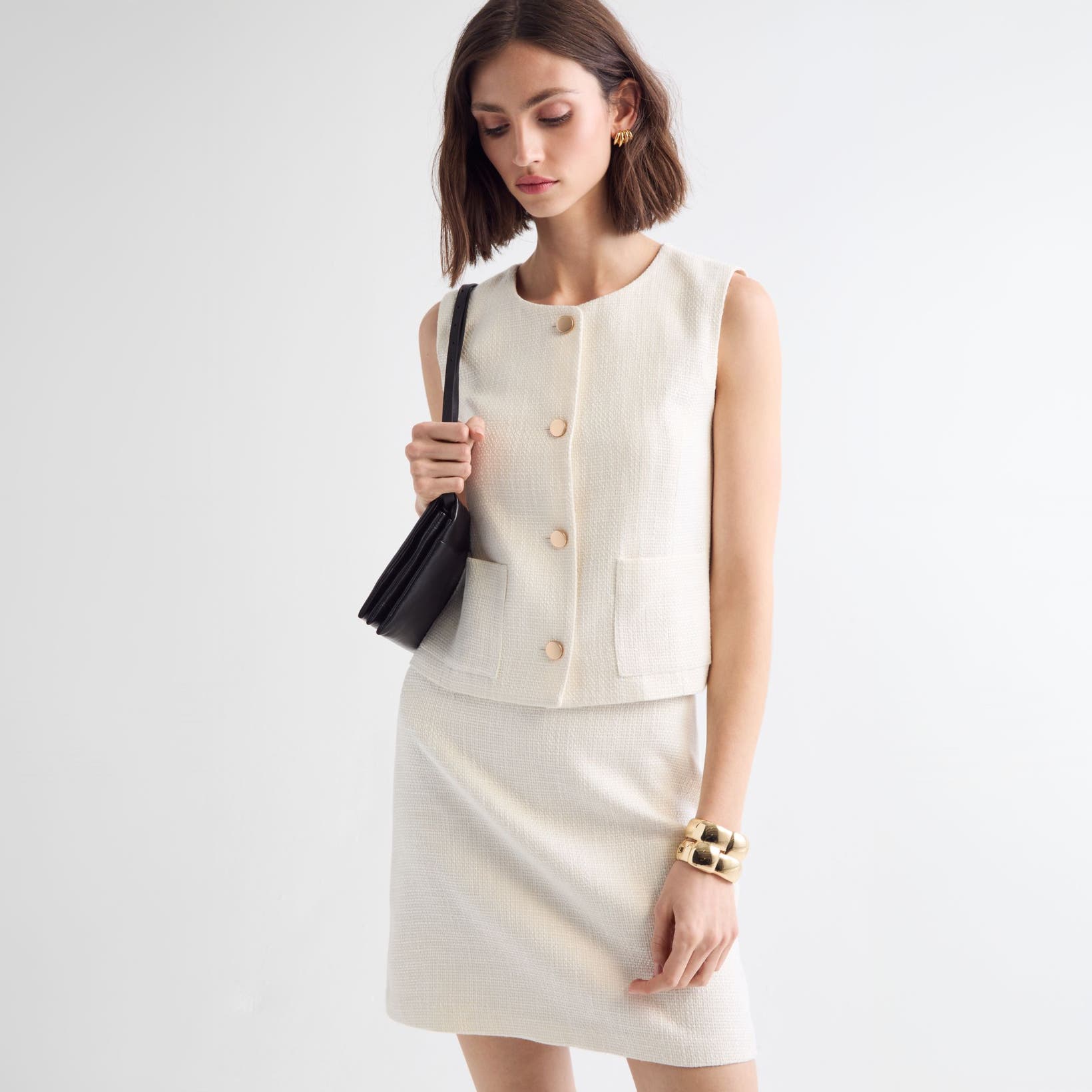 Woman wearing a white vest and skirt with a handbag and jewelry.