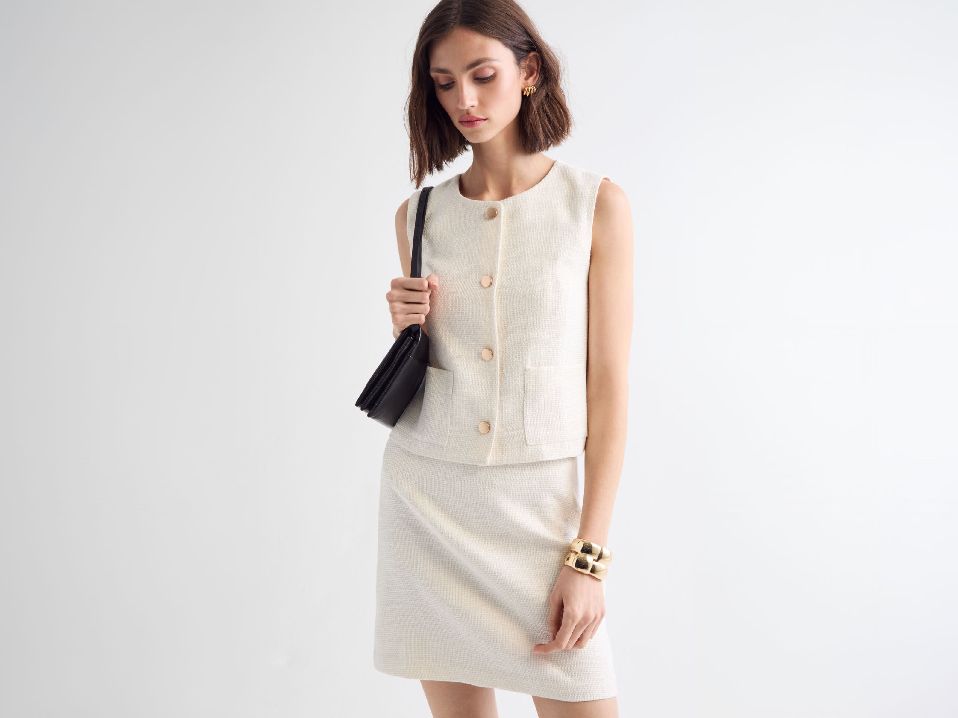 Woman wearing a white vest and skirt with a handbag and jewelry.