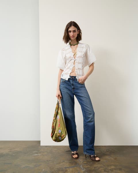 A woman wearing a tie-front lace top, jeans and sandals with a handbag.