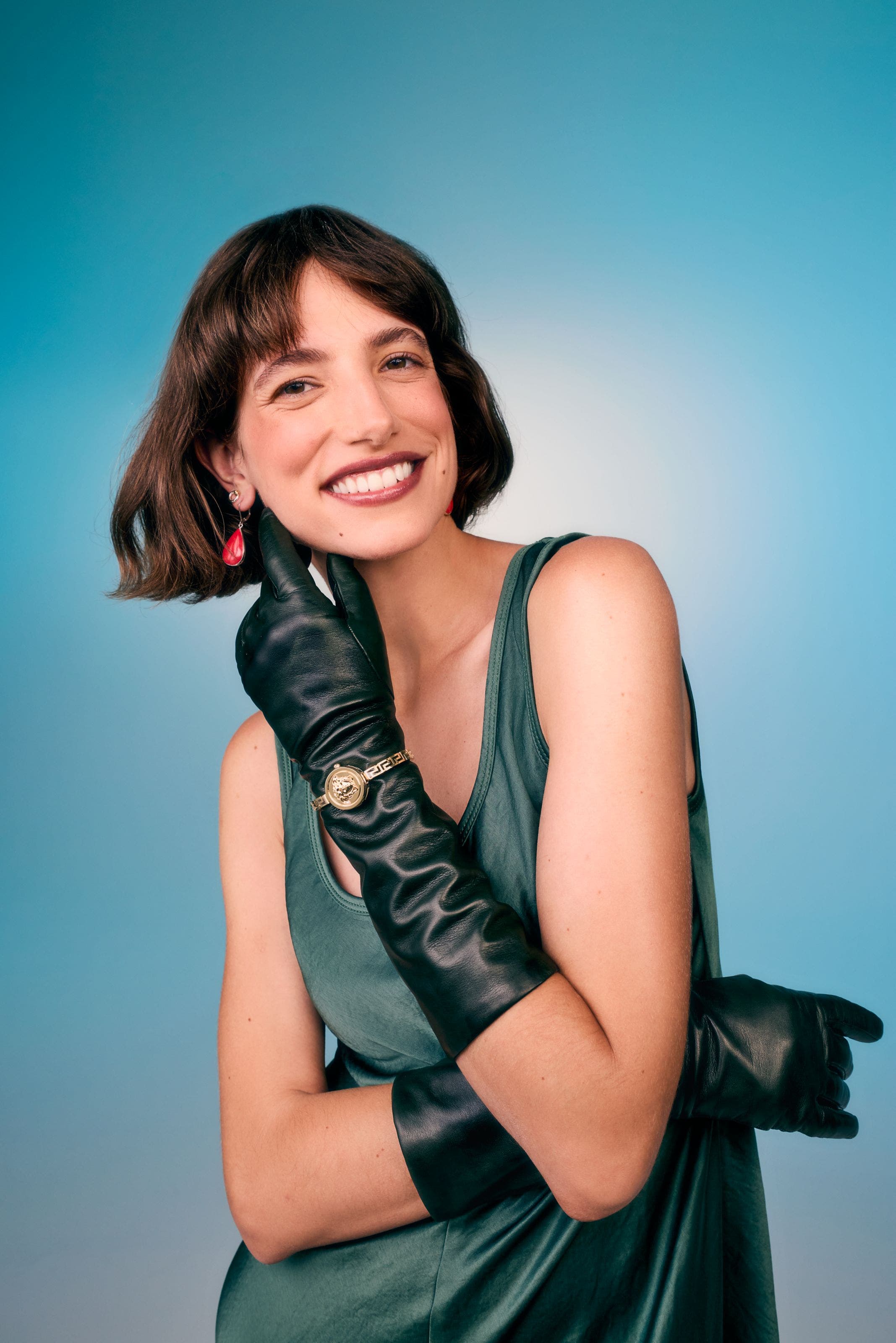 Woman in a green sleeveless top, wearing long black leather gloves with a gold watch over one, touching a red drop earring.