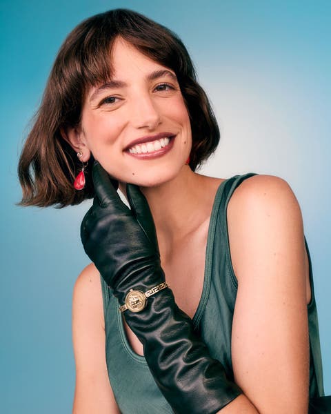 Woman in a green sleeveless top, wearing long black leather gloves with a gold watch over one, touching a red drop earring.