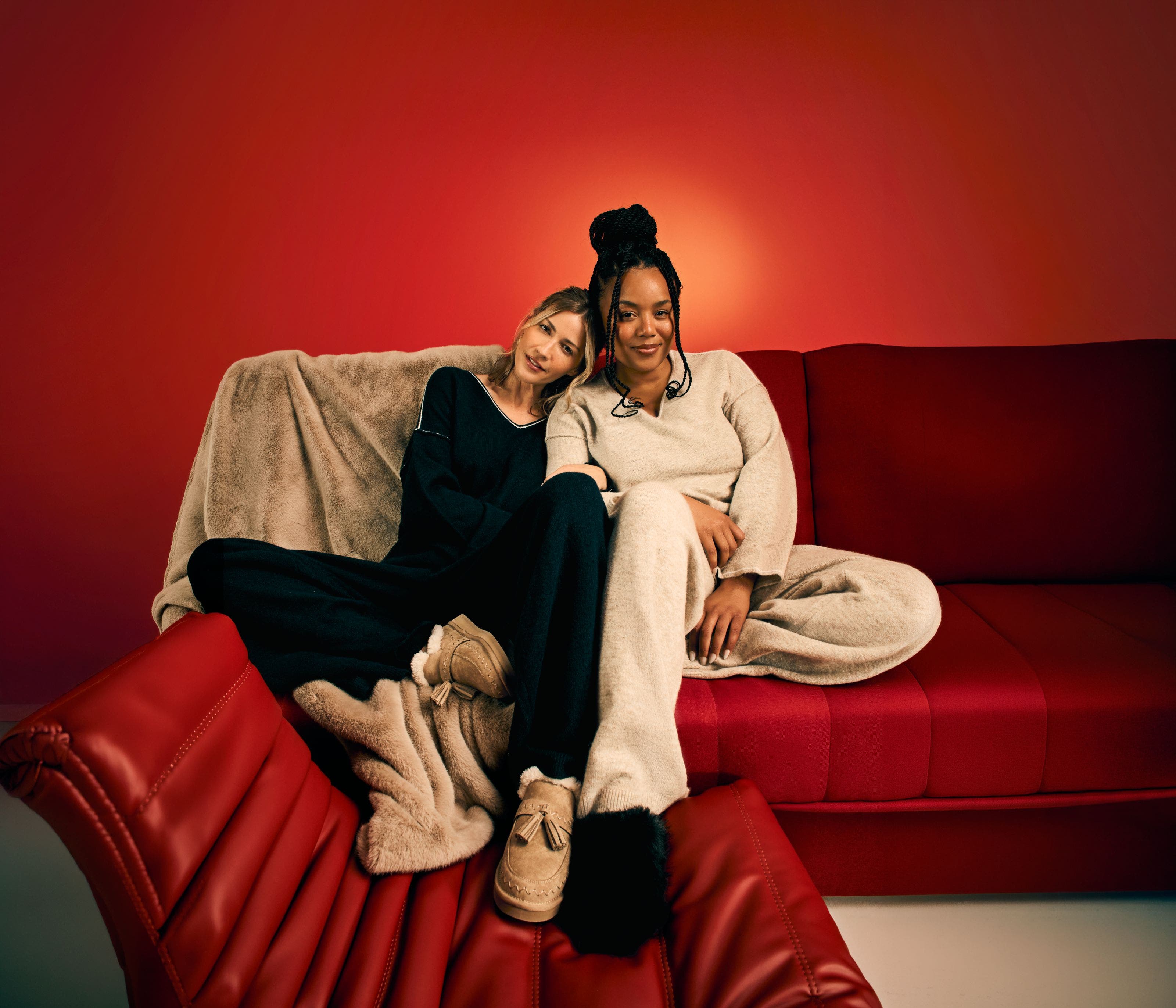 Two women wearing loungewear relax on a couch.
