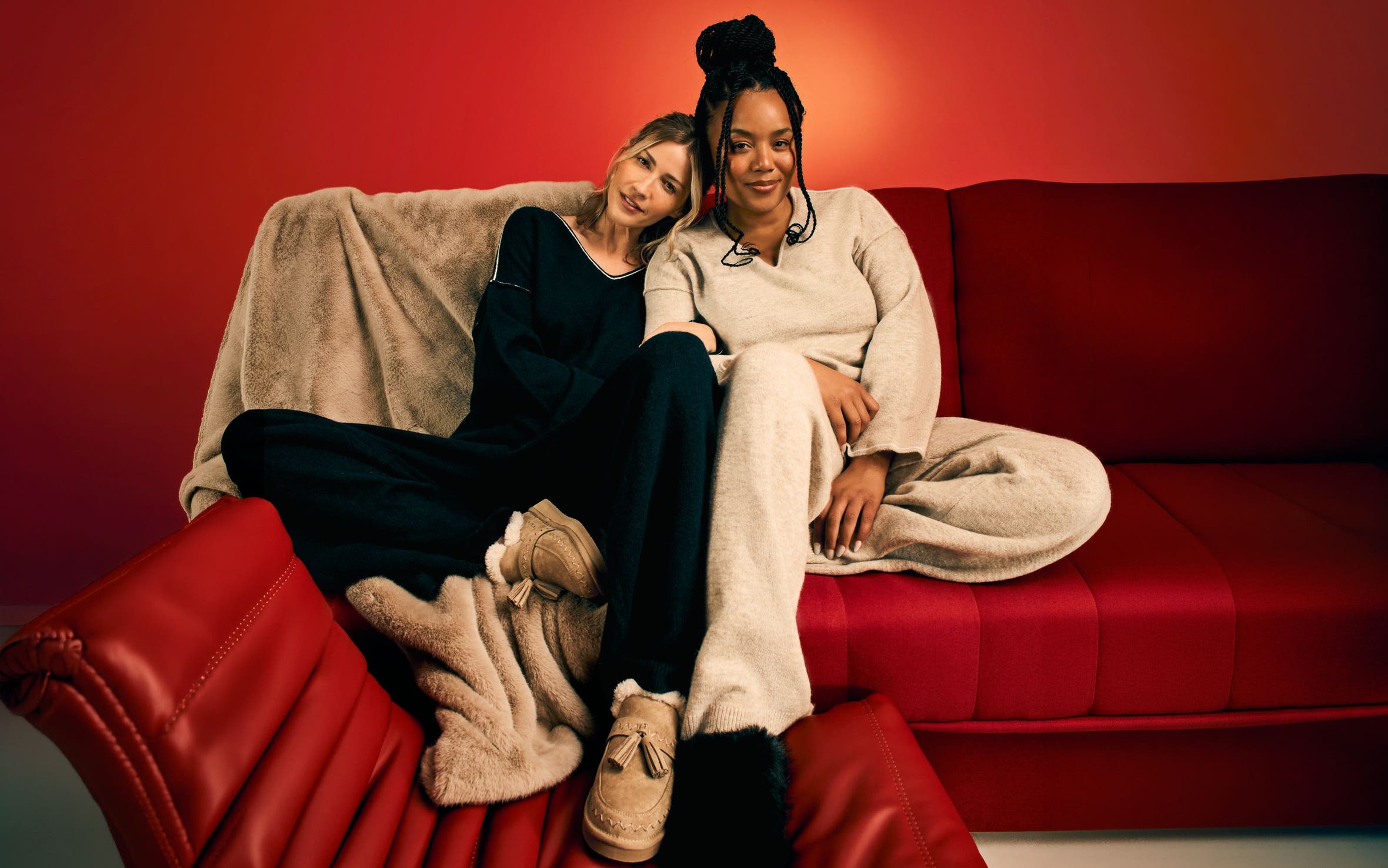 Two women wearing loungewear relax on a couch.