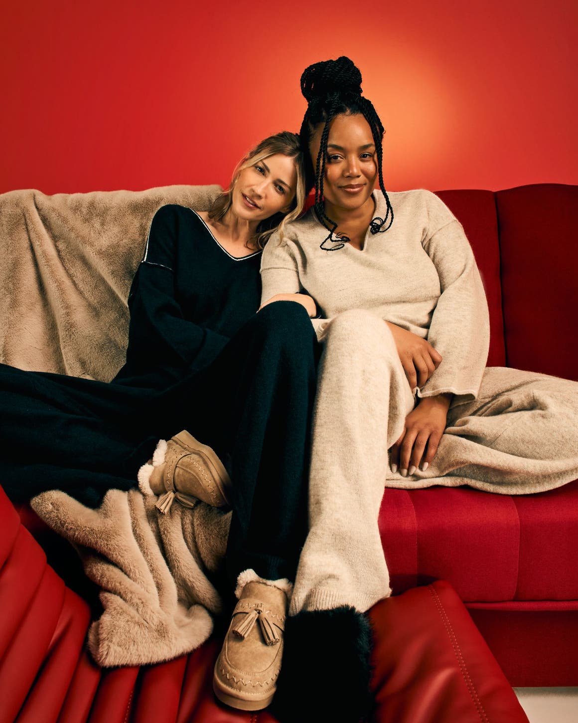 Two women wearing loungewear relax on a couch.