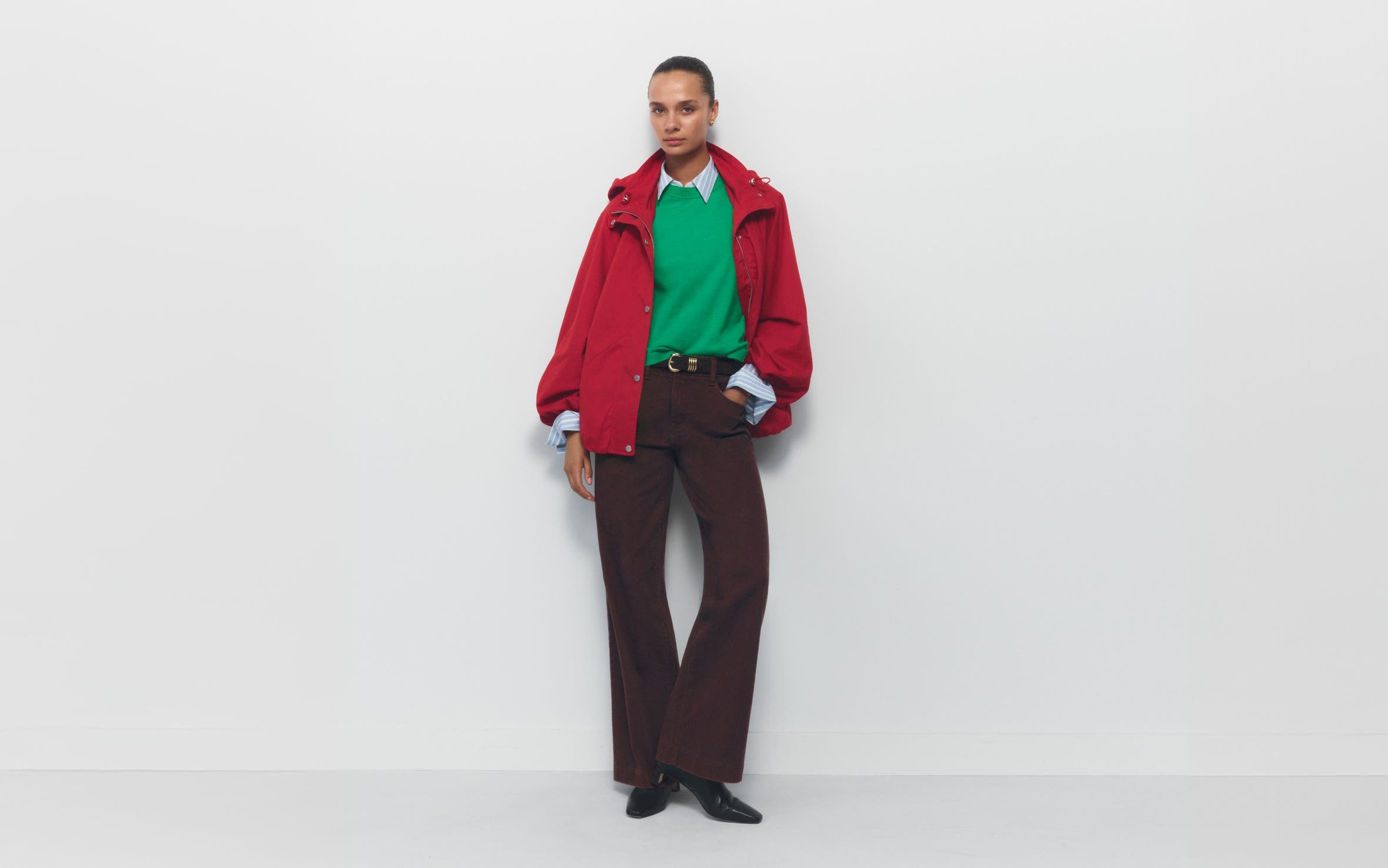 A woman wearing a green sweater and red coat over a button-up with pants and boots.