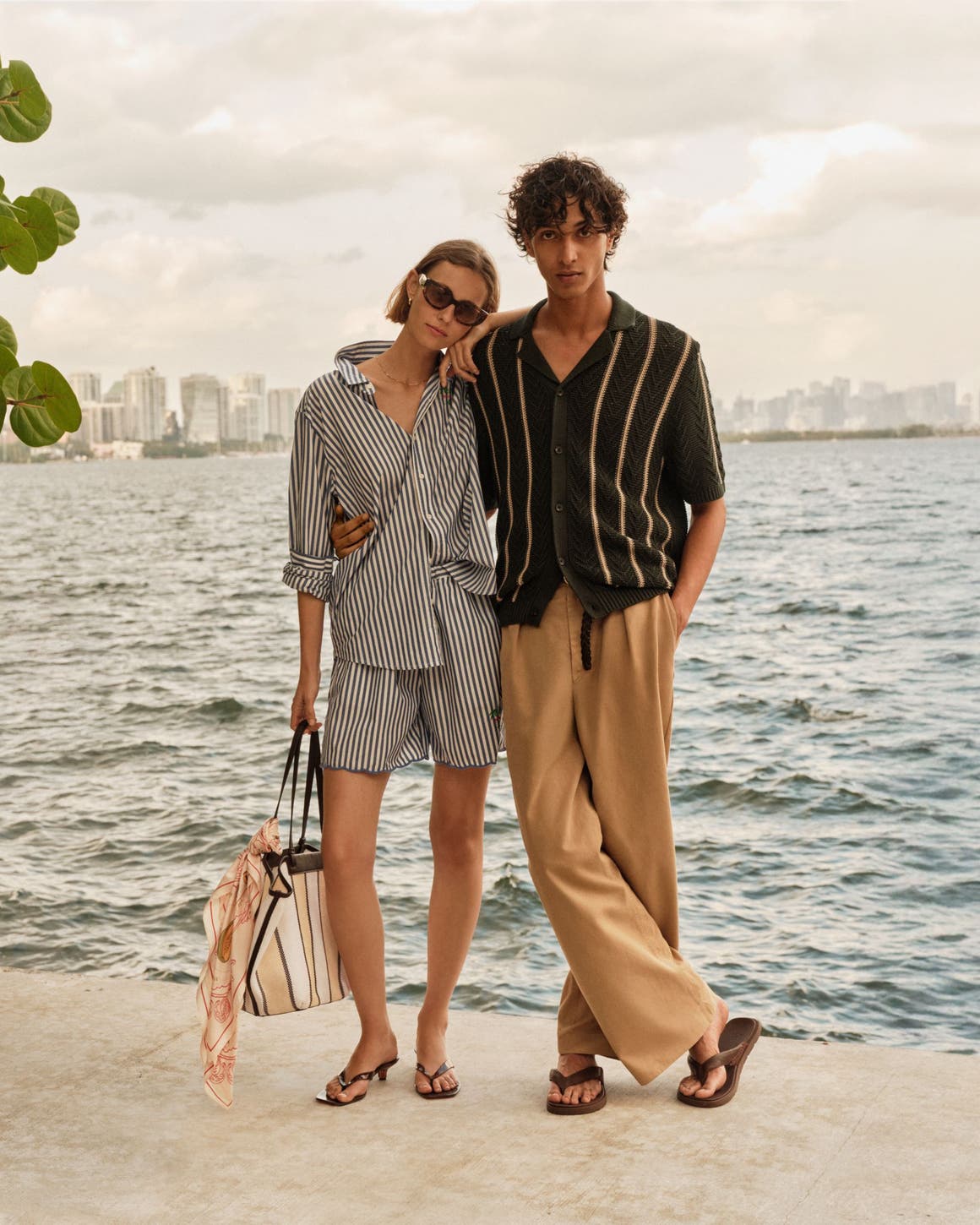 A man and woman in summer travel outfits by a lake with a city skyline in the distance. 