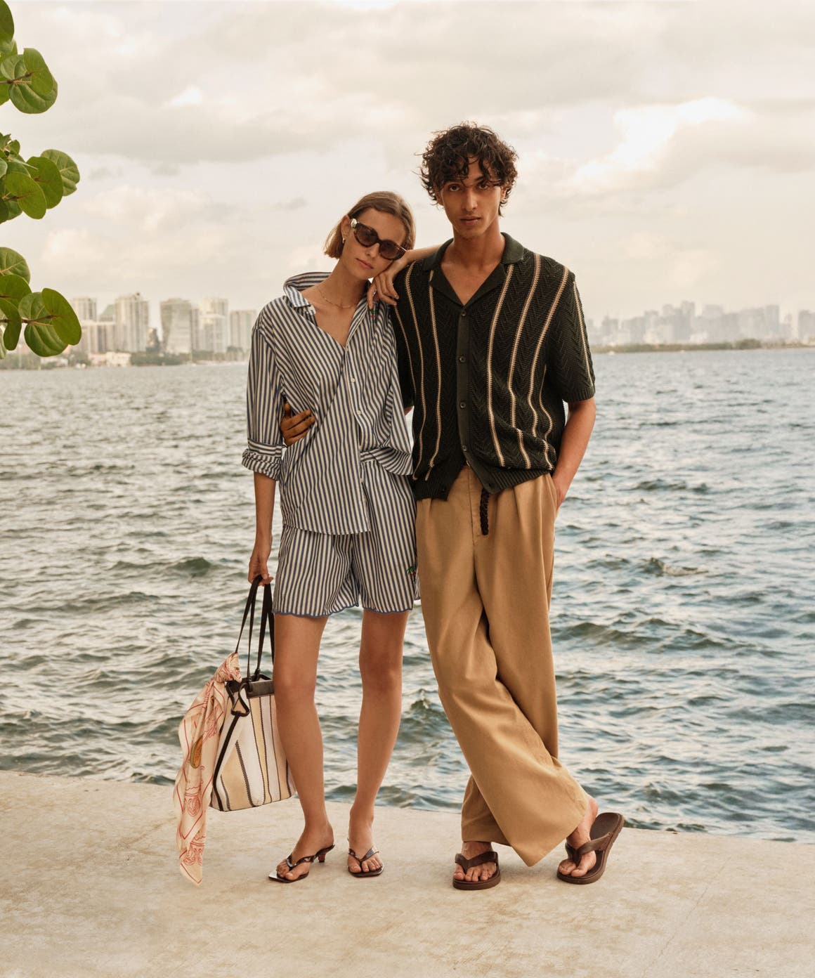 A man and woman in summer travel outfits by a lake with a city skyline in the distance. 