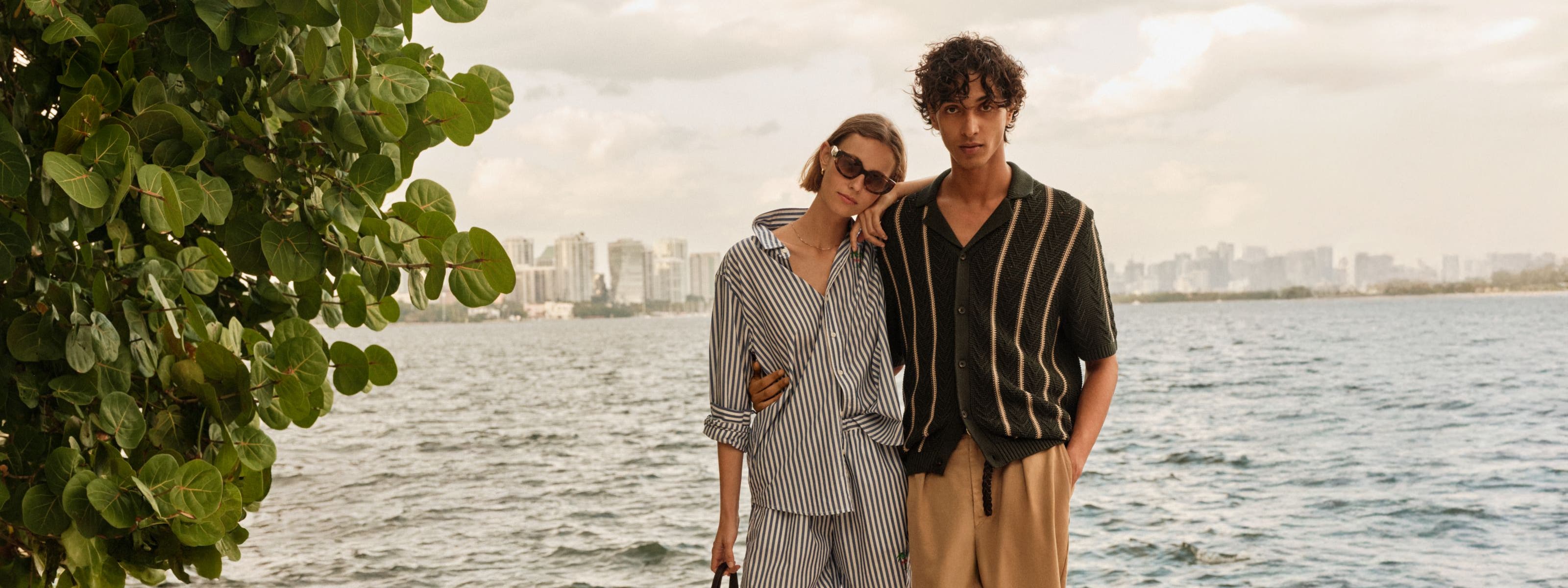 A man and woman in summer travel outfits by a lake with a city skyline in the distance. 