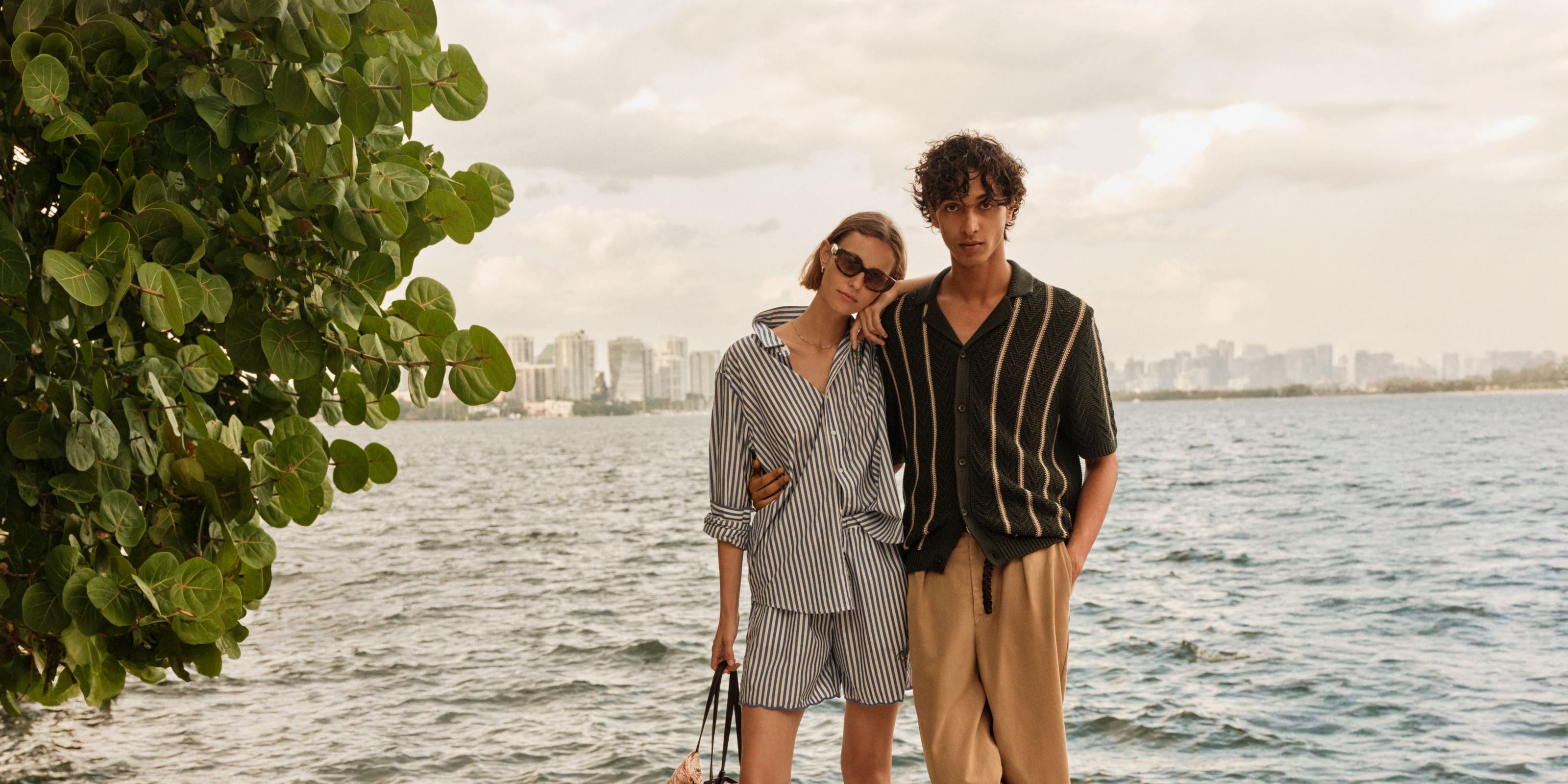 A man and woman in summer travel outfits by a lake with a city skyline in the distance. 