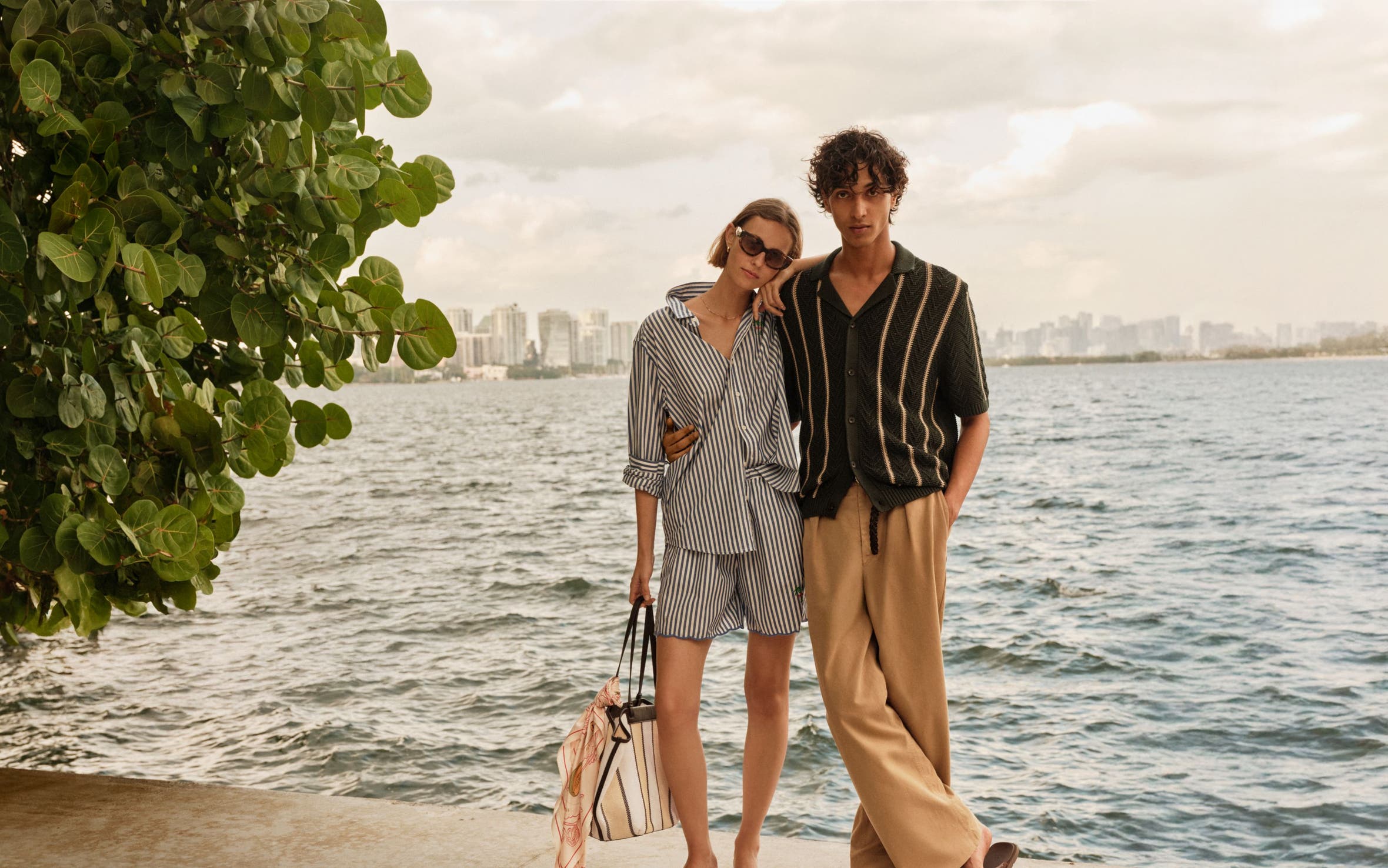 A man and woman in summer travel outfits by a lake with a city skyline in the distance. 