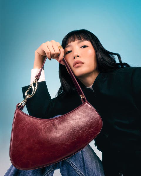 Model holding a burgundy crescent-shaped shoulder bag with gold chain detail.