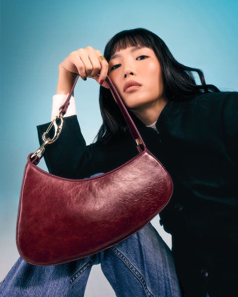 Model holding a burgundy crescent-shaped shoulder bag with gold chain detail.
