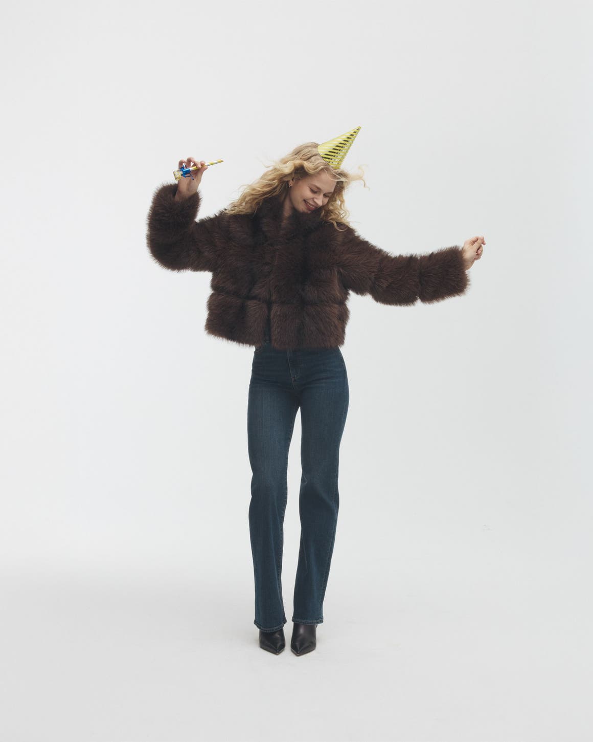 Woman shimmying in a faux-fur jacket, jeans and party hat.