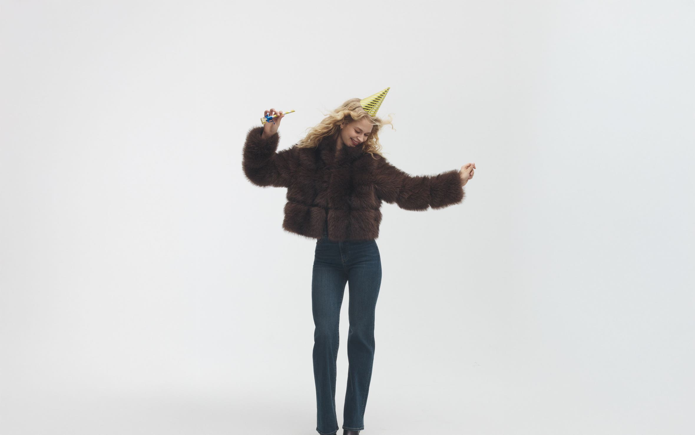 Woman shimmying in a faux-fur jacket, jeans and party hat.