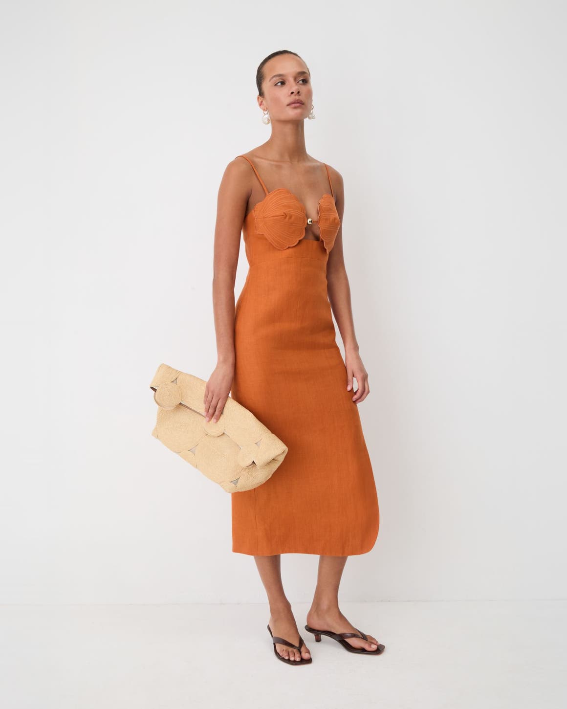 Woman wearing a oranage terra cotta sleeveless midi shell dress, with a Cult Gaia clutch.