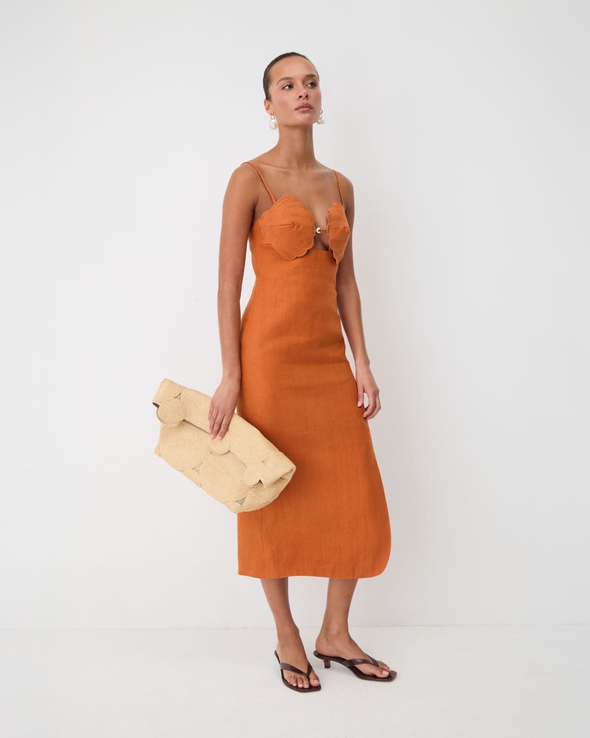 Woman wearing a oranage terra cotta sleeveless midi shell dress, with a Cult Gaia clutch.