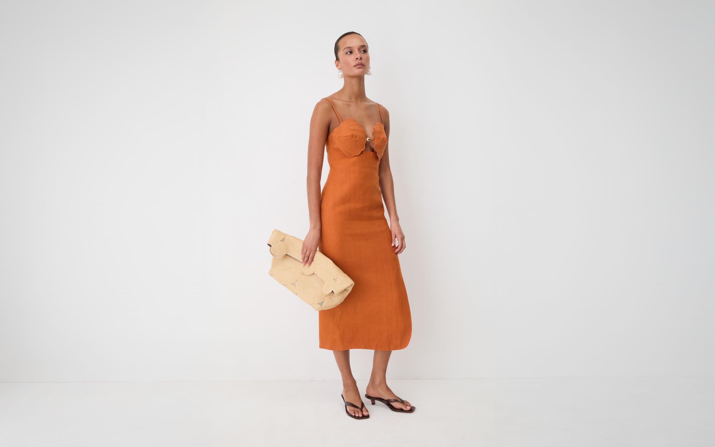 Woman wearing a oranage terra cotta sleeveless midi shell dress, with a Cult Gaia clutch.