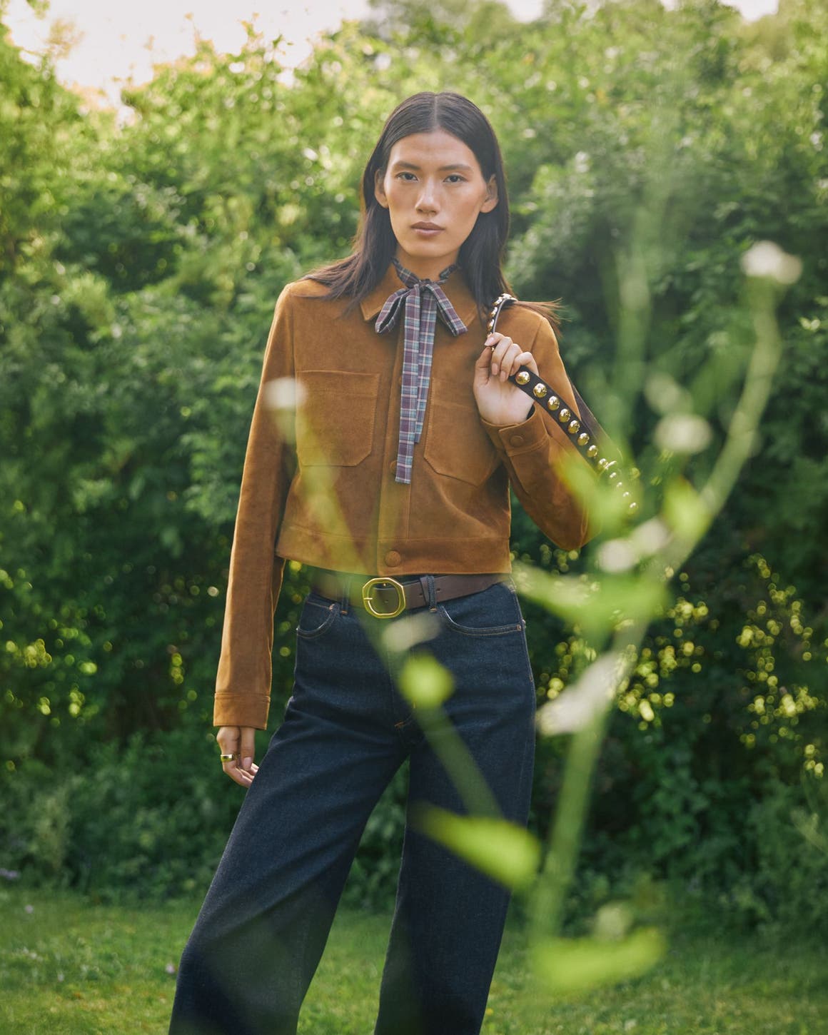 A woman wearing a tan suede jacket and dark denim jeans stands in a lush green garden.